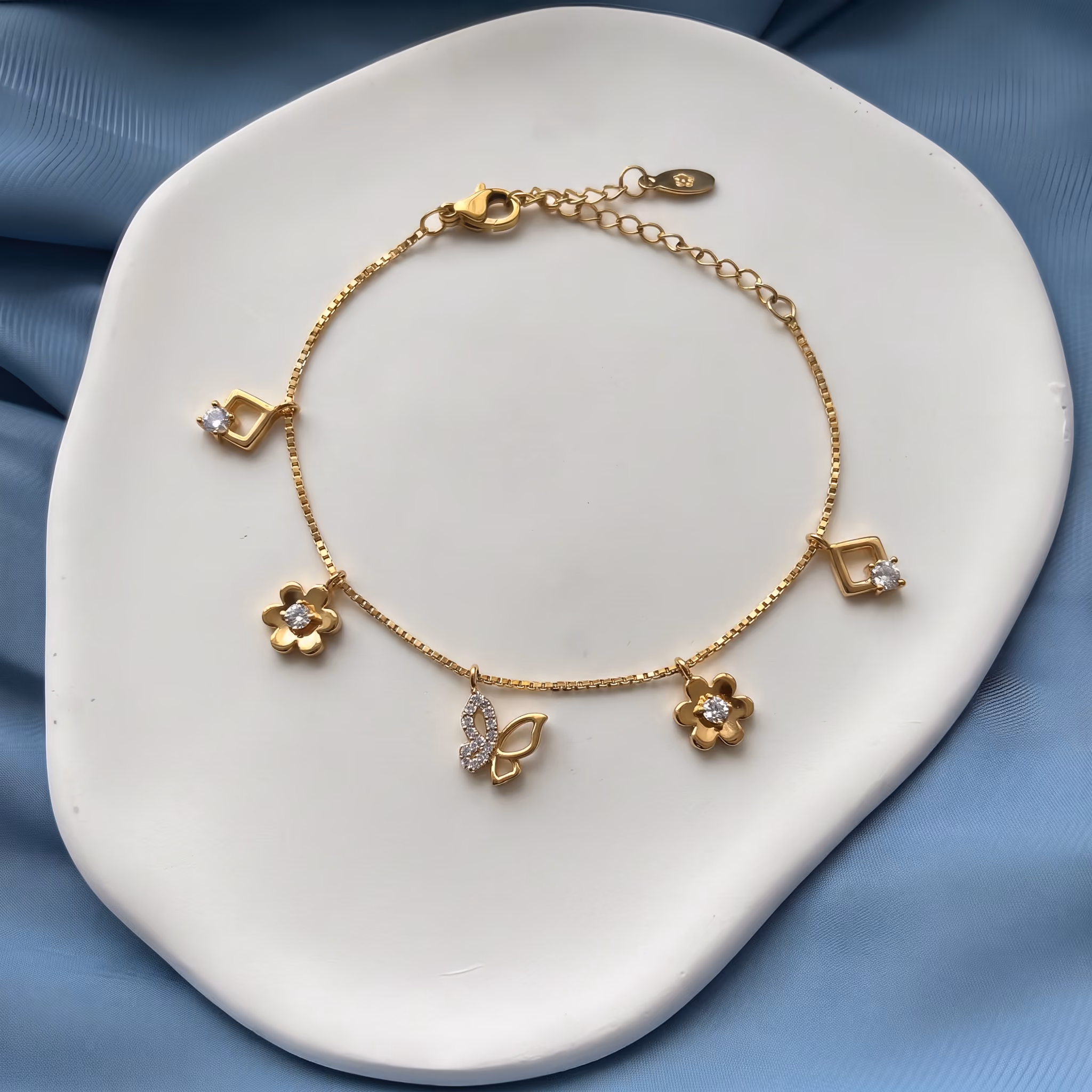 Gold bracelet with flower, butterfly, and diamond-shaped charms on a white dish with blue fabric.