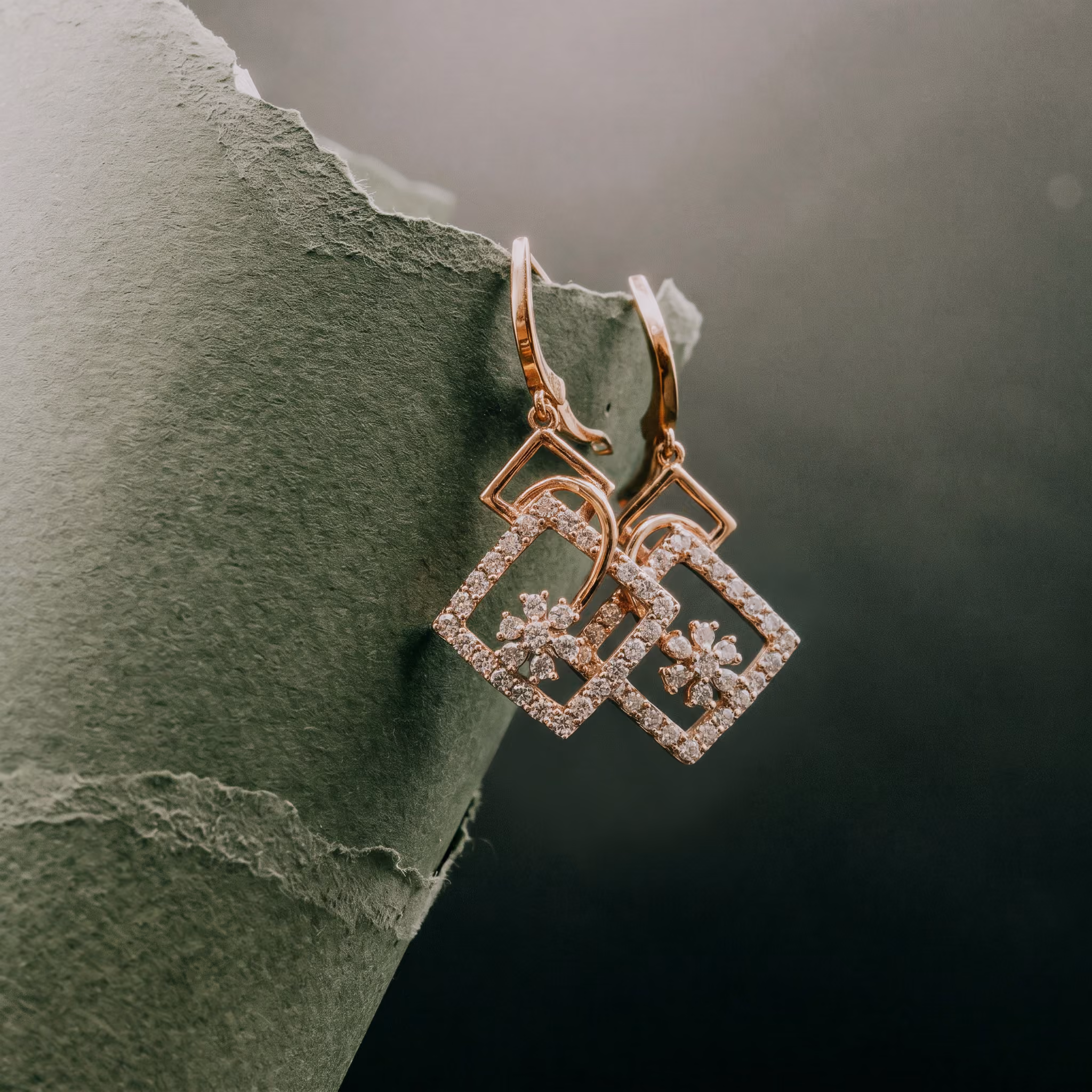 Pair of gold square earrings with diamonds and floral design hanging on torn green paper.