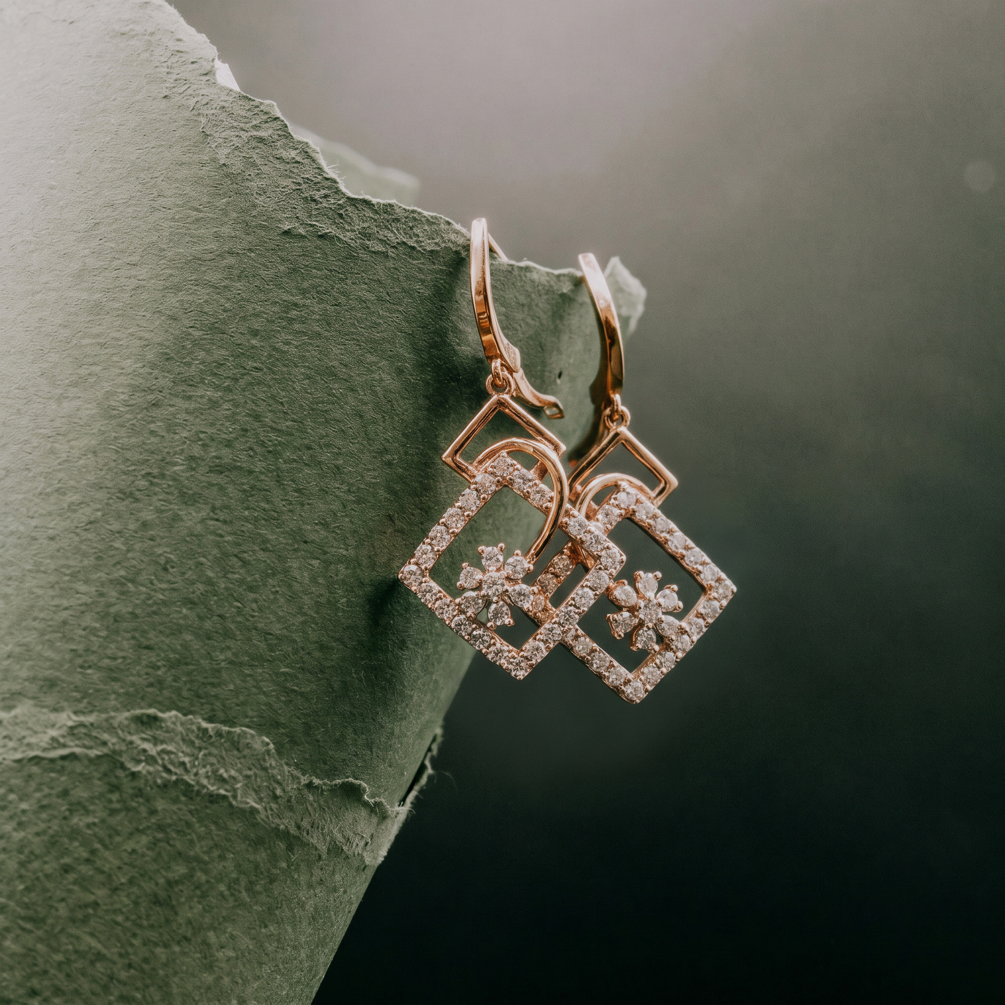 Pair of gold square earrings with diamonds and floral design hanging on torn green paper.