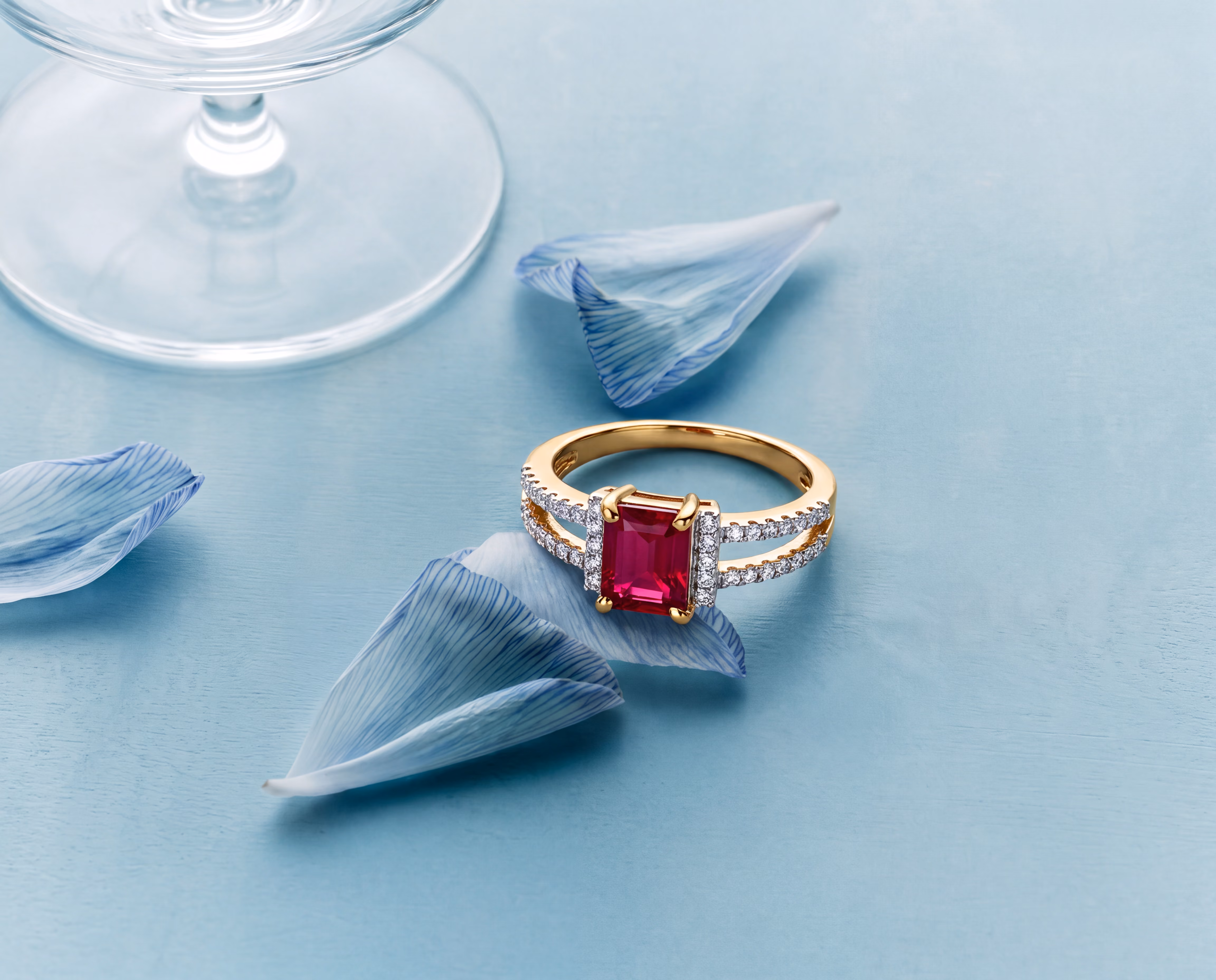 Gold ring with rectangular red ruby and diamond accents placed among blue flower petals on light blue surface.