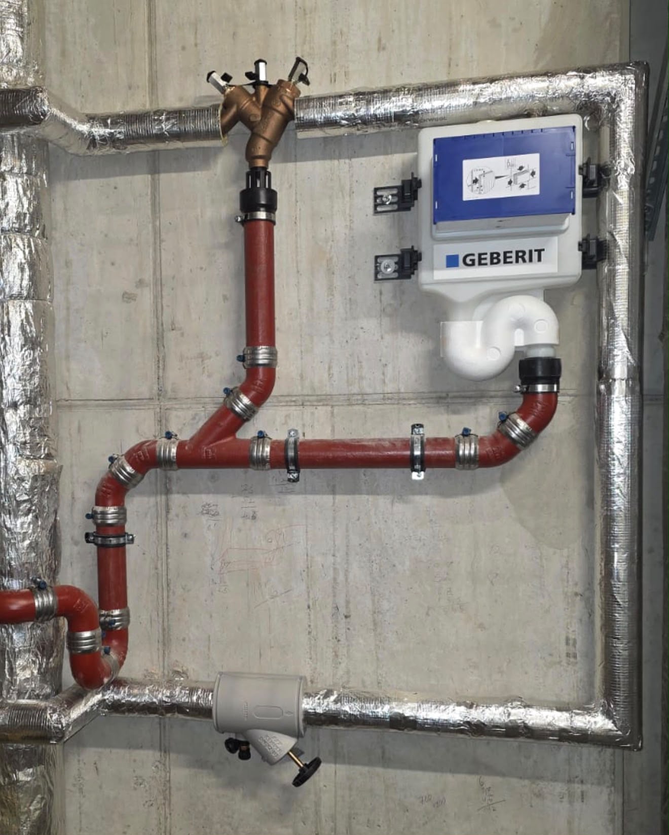 Rotes Rohrleitungssystem mit Geberit-Montagekasten und isolierten Rohren an Betonwand.
