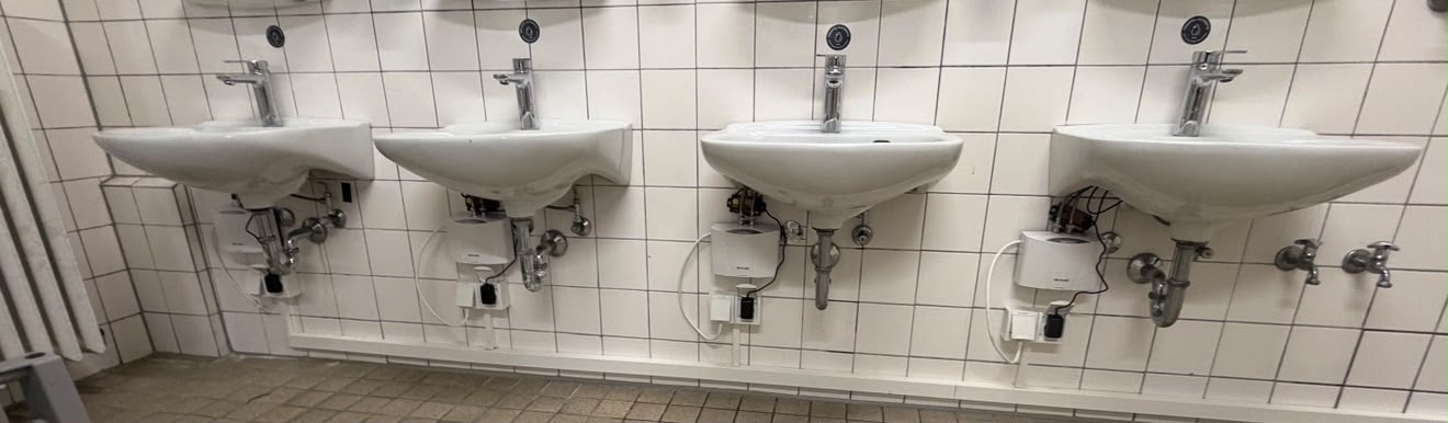 Vier weiße Waschtische mit Wasserhähnen, Fliesenwand und Boden in einem Badezimmer.