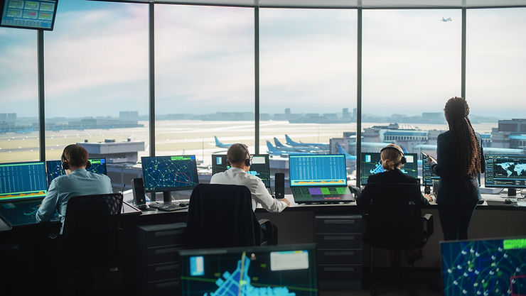 Air traffic controllers in a tower monitor multiple screens. View of airport and planes outside. Calm, focused atmosphere. Showing how secured communication is important