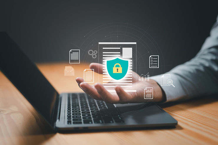 Hand holding digital documents with a security shield icon above a laptop on a wooden table, symbolizing data protection and cybersecurity.