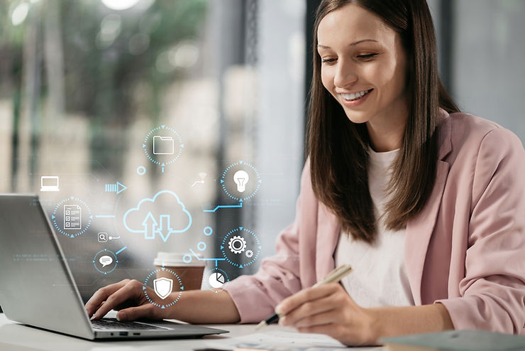 Smiling woman in a pink blazer at a desk using a laptop and writing, with holographic cloud icons. Office setting with a bright, modern feel.