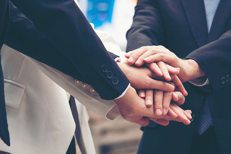 Hands stacked in a team gesture, individuals in suits. The setting is likely professional, conveying unity and collaboration.