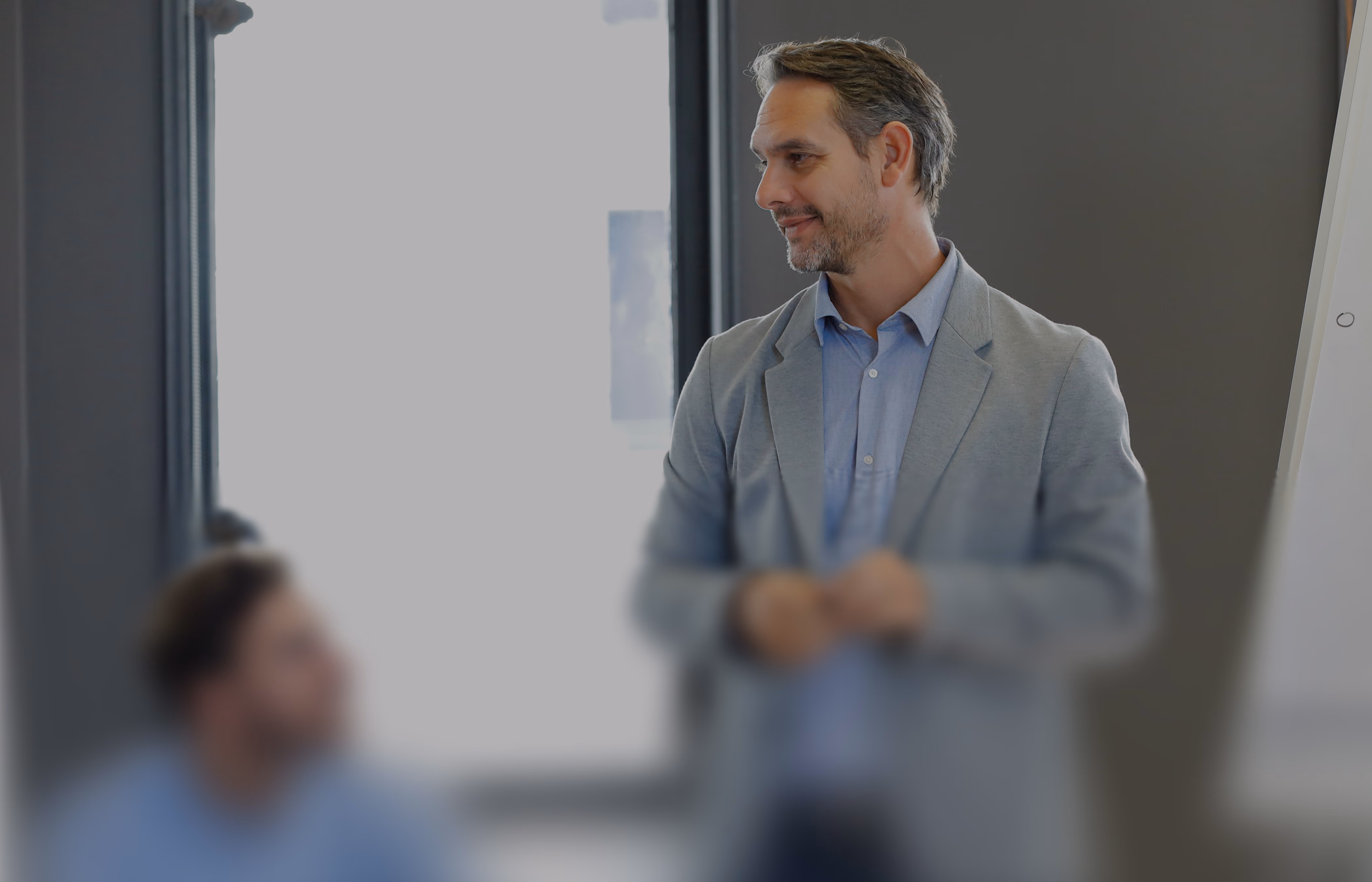 Man in gray blazer smiling and speaking to a seated person in an office with a whiteboard.
