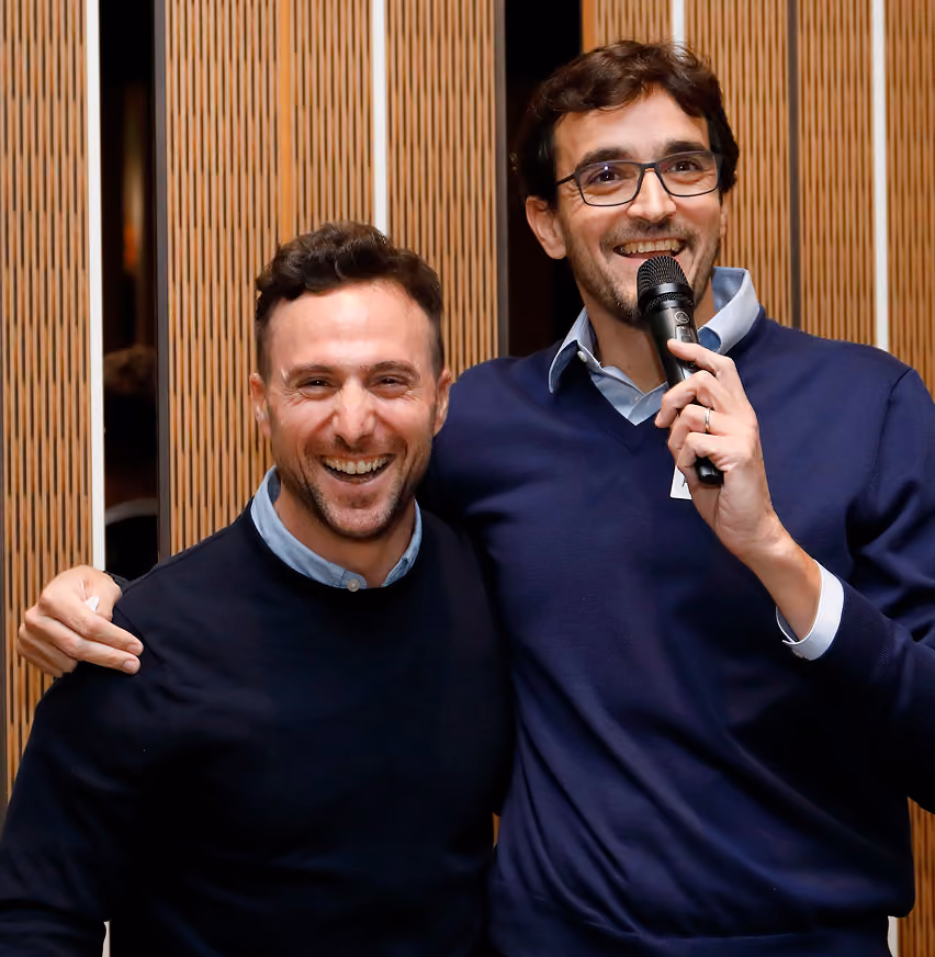 Two men smiling, one with glasses speaking into a microphone, standing close in front of wooden panels.