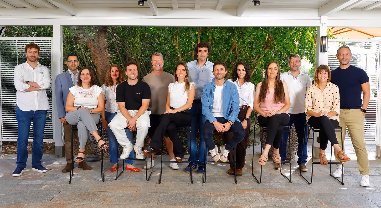 Group of fifteen adults smiling in casual clothing in a covered outdoor space with greenery behind them.