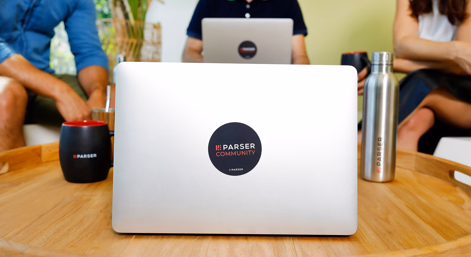 Laptop with Parser Community sticker on a wooden table surrounded by two people and branded mugs and bottle.