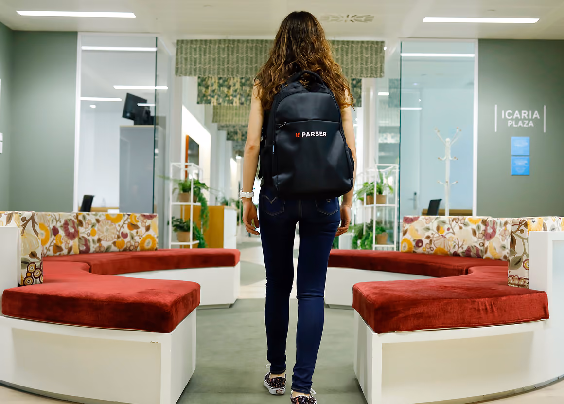 Person wearing a Parser backpack walking through a bright office lobby with red sofas.