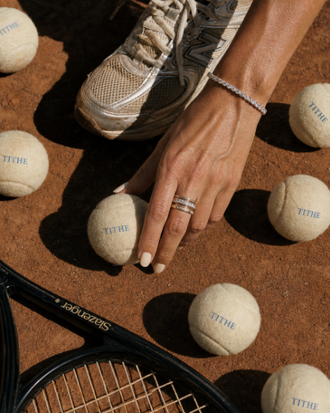 A hand placing TITHE tennis balls on a clay court.
