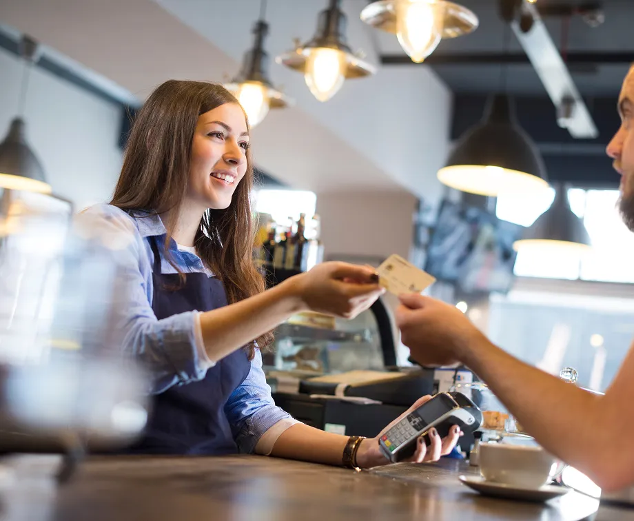Smiling barista accepts a credit card payment at a cafe counter with coffee cup nearby.