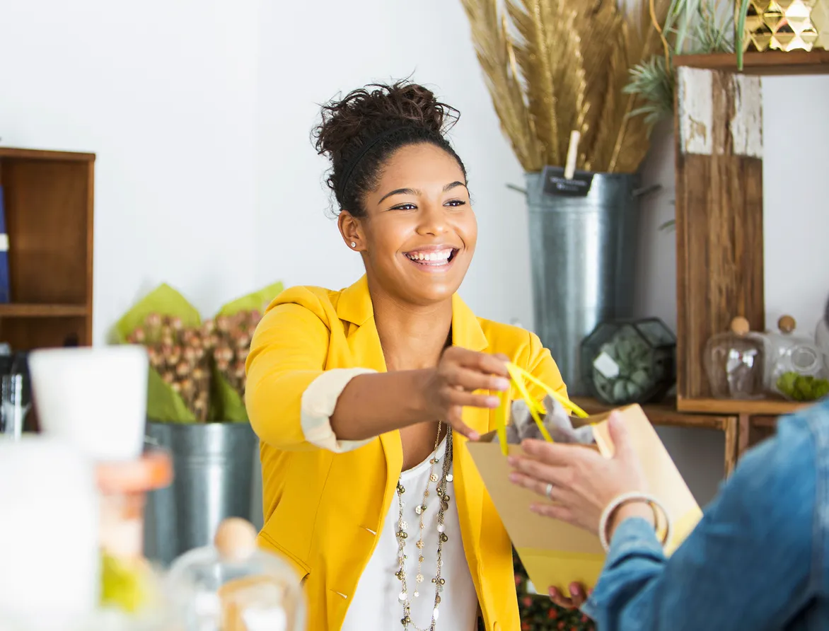 Smiling woman in yellow blazer hands over a small shopping bag in a cozy shop.