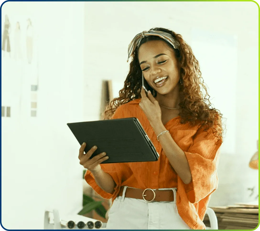 Smiling woman in orange blouse speaking on phone and holding a tablet device indoors.