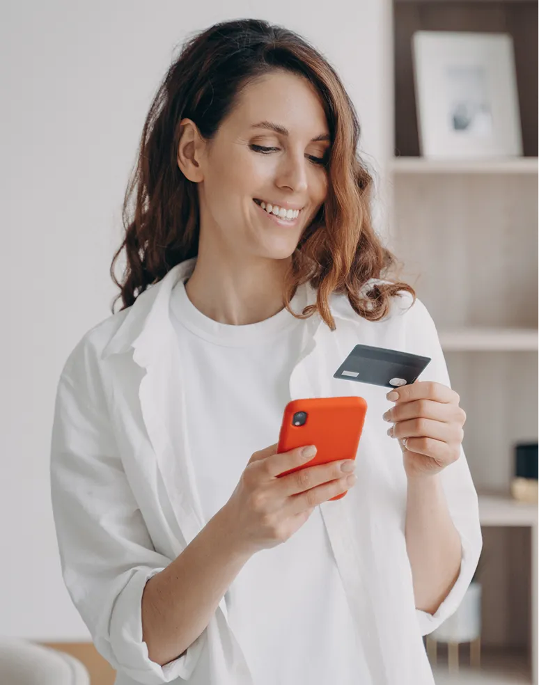 Smiling woman holding a credit card and orange smartphone indoors, looking at the card.