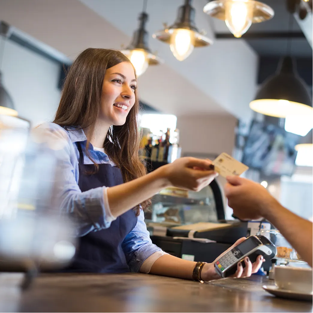 Smiling cashier holding a card reader as customer hands over a credit card at a counter.