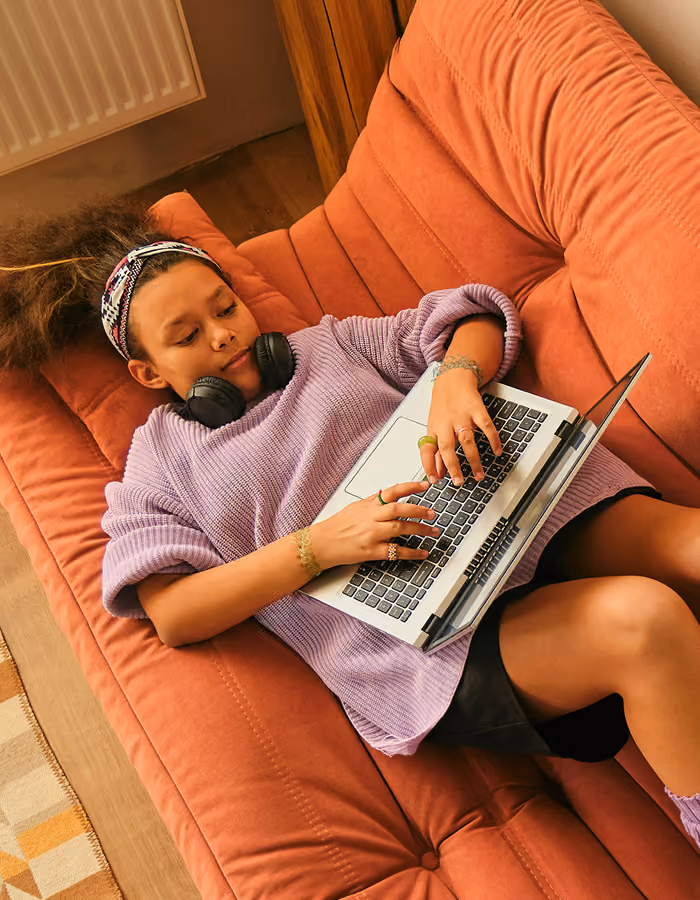 Young person in purple sweater lying on rust-colored couch using a laptop with headphones around neck.