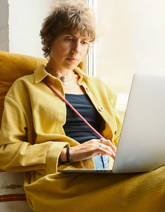 Young person in purple sweater lying on rust-colored couch using a laptop with headphones around neck.