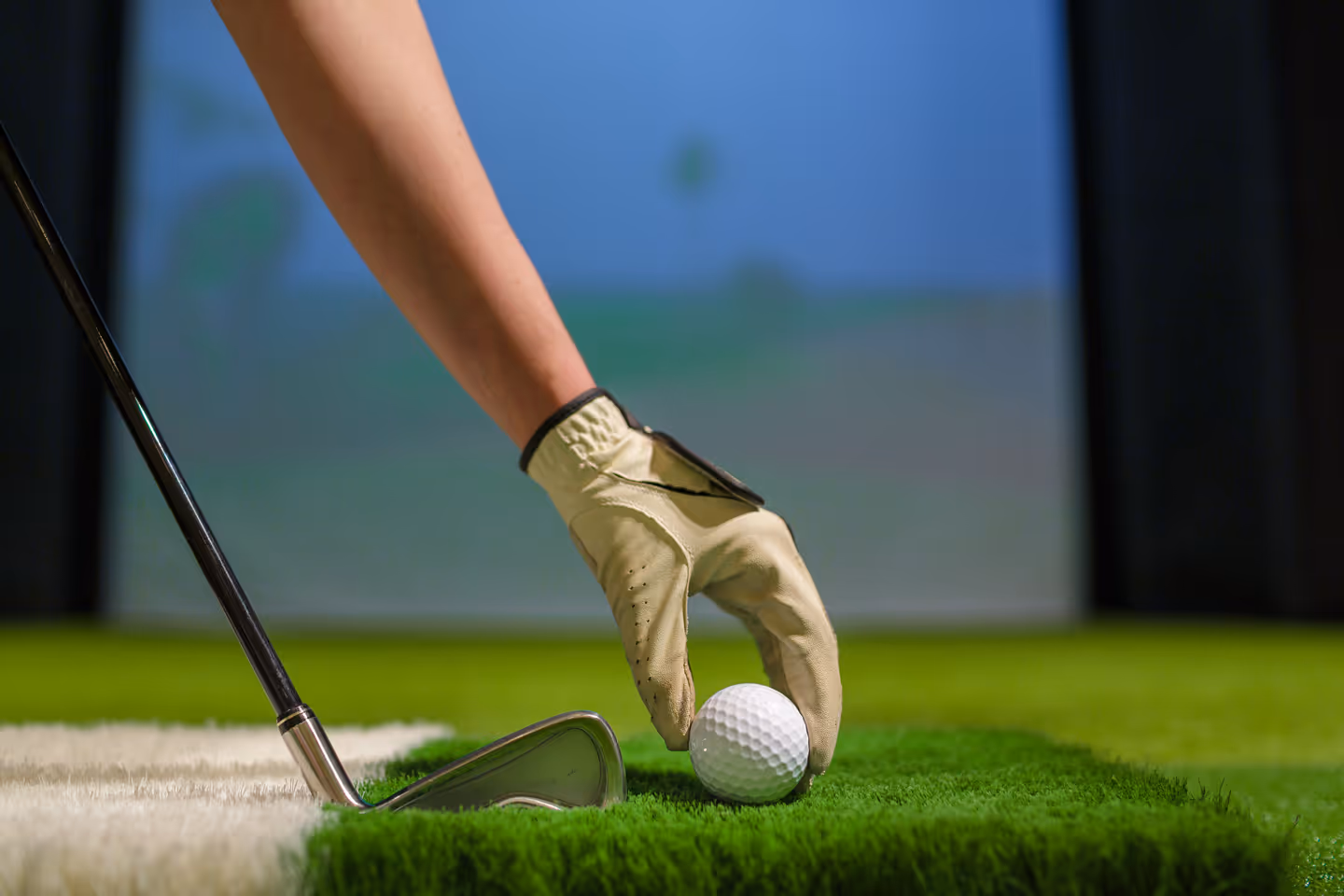 Hand with golf glove placing a golf ball on artificial grass next to a golf iron club.