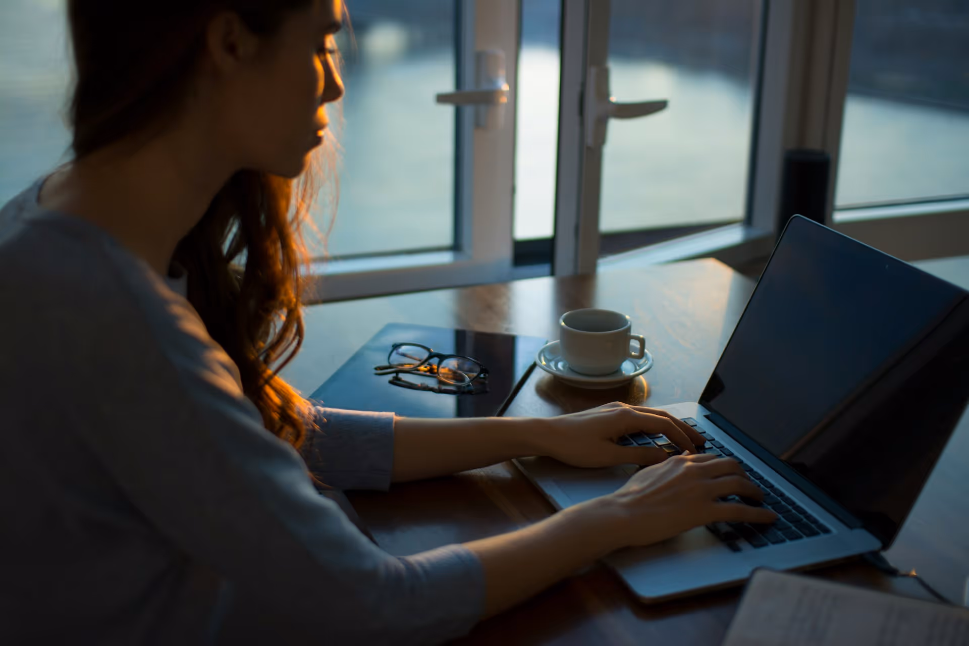 Woman typing on laptop near window with a cup and glasses on the table at sunset.