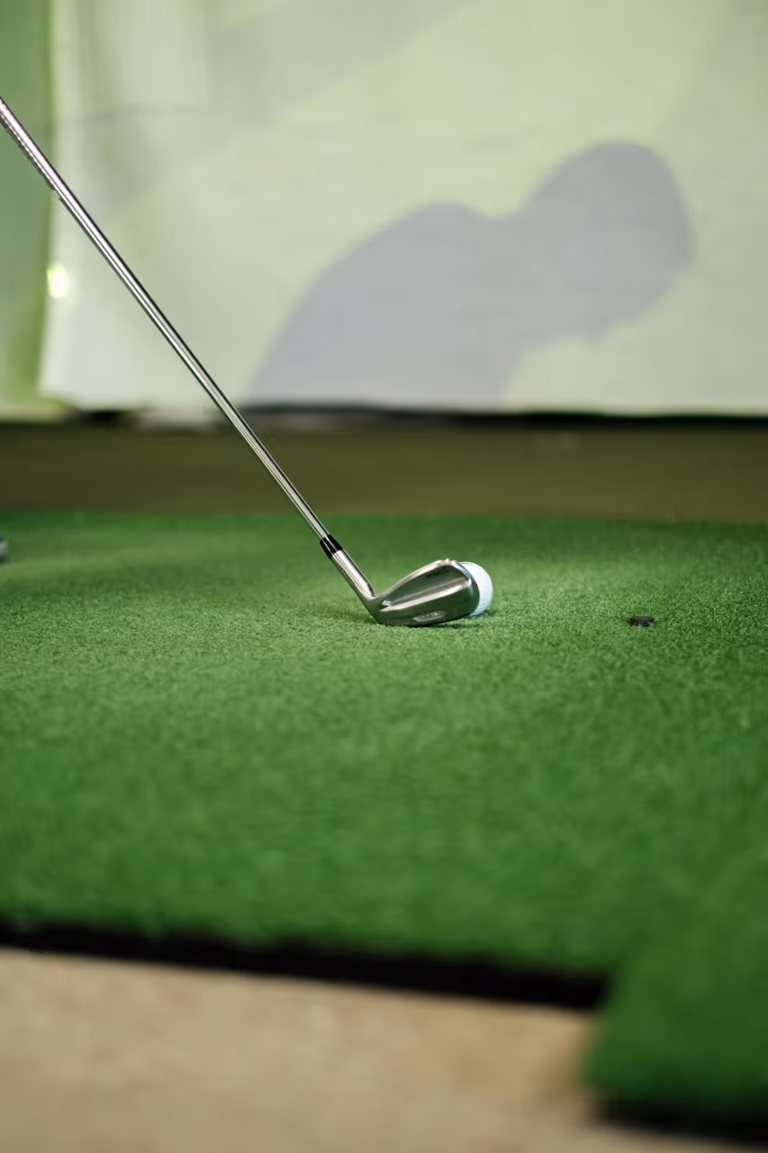 Golf club positioned behind a golf ball on green turf indoors with a shadow in the background.