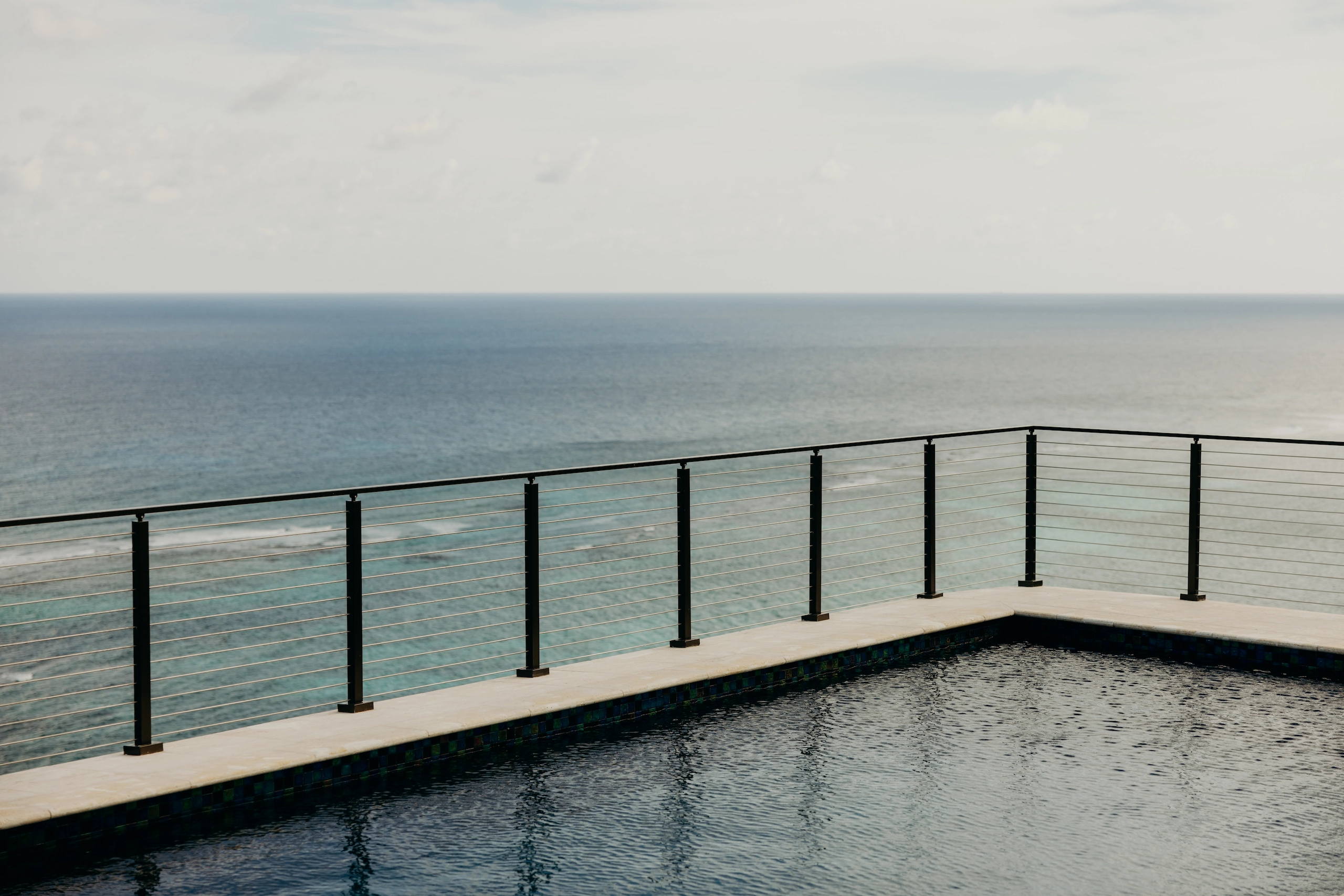 Pool with metal cable railing overlooking calm ocean under cloudy sky.