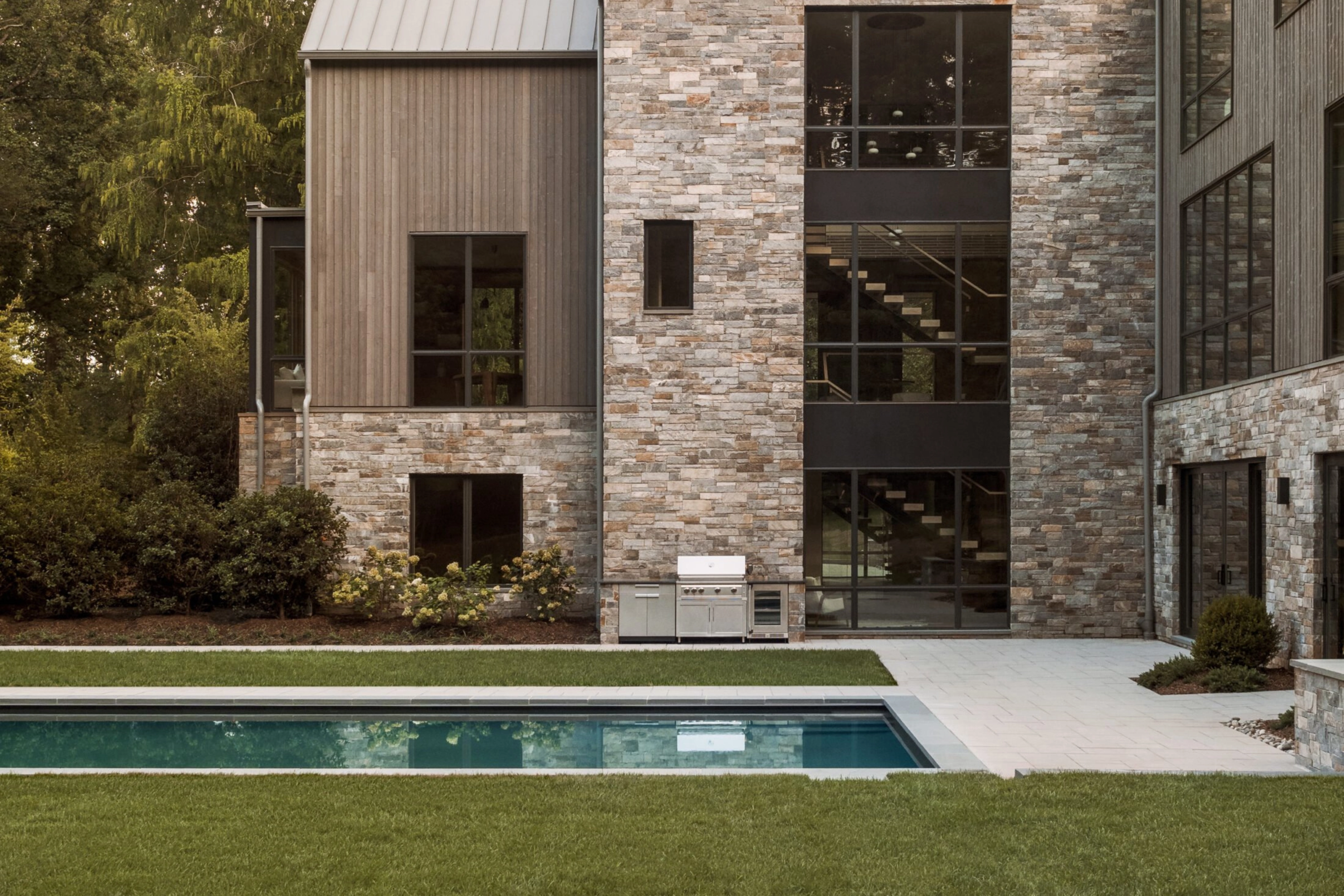 Modern stone and wood house exterior with large windows showcasing a FLIGHT Mono staircase inside, with a rectangular pool in front.
