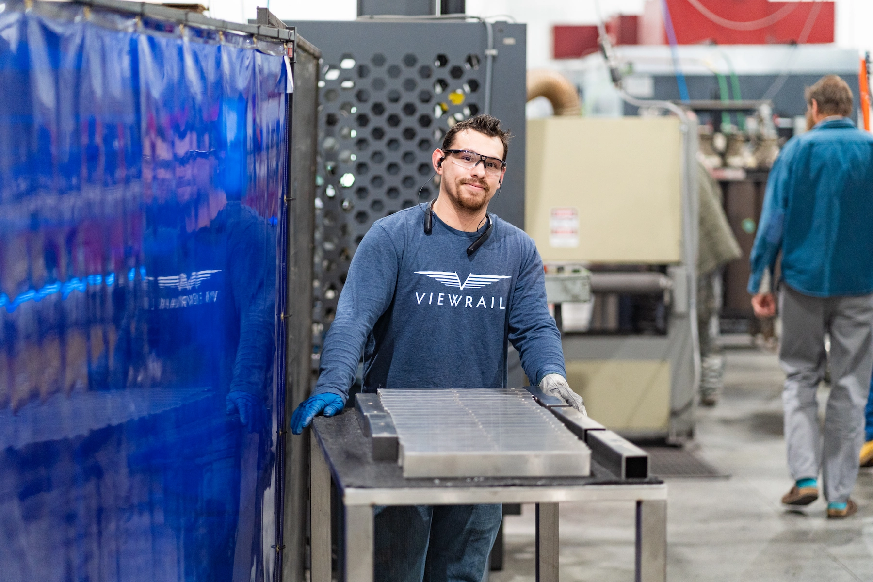Viewrail factory worker with Viewrail shirt wearing gloves and safety glasses standing behind a metal table with metal parts.