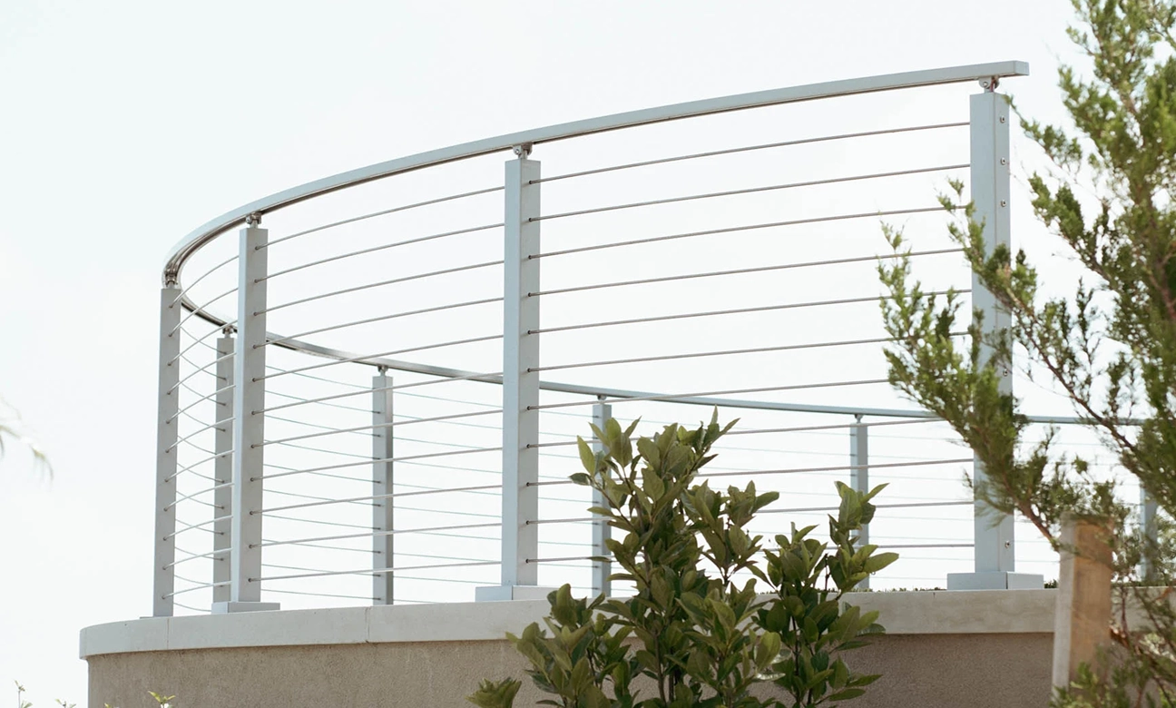 Curved stainless steel cable railing on a beige concrete platform with green plants nearby.