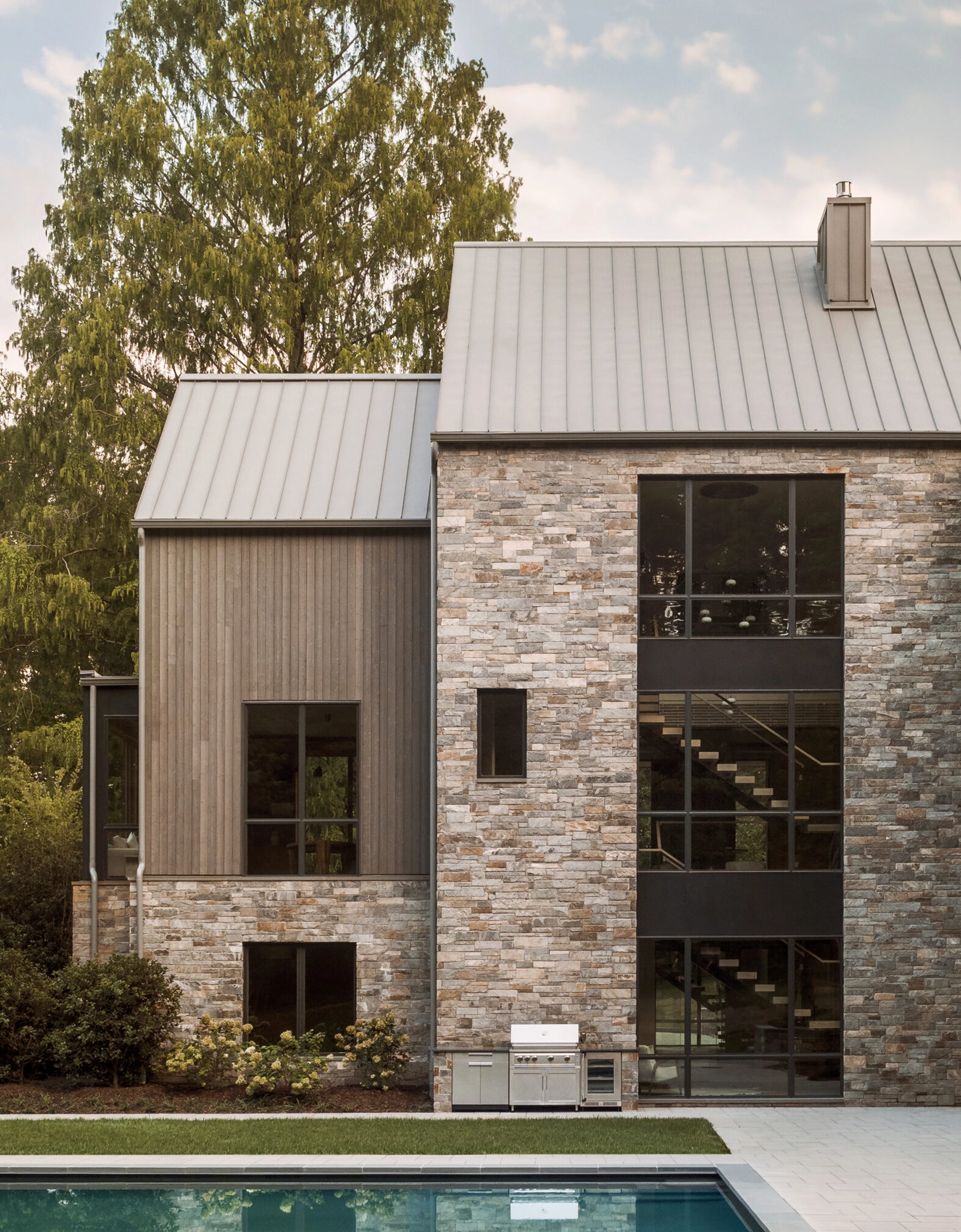 Modern stone and wood house exterior with a FLIGHT Mono system visible through large windows, metal roof, and pool in front.