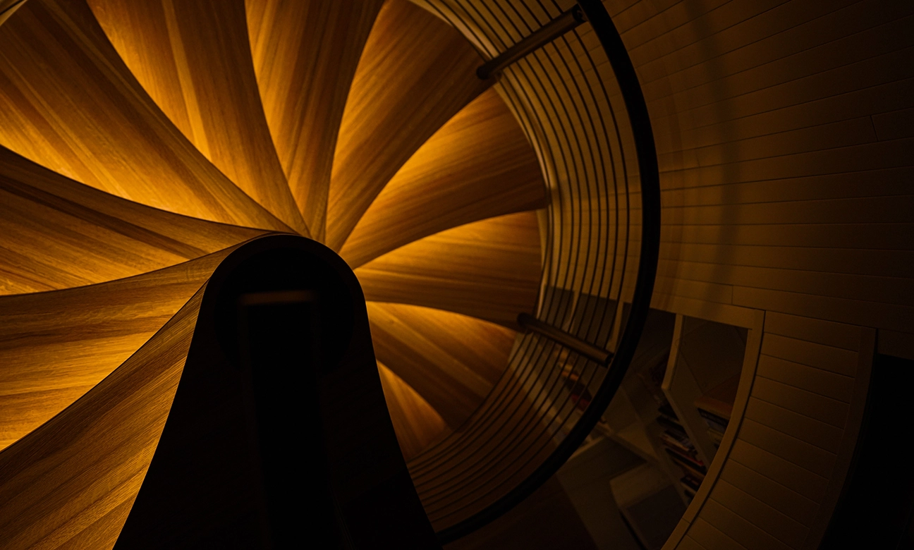 Top-down view of a warmly lit wooden spiral staircase with black metal railing and wall shelves.