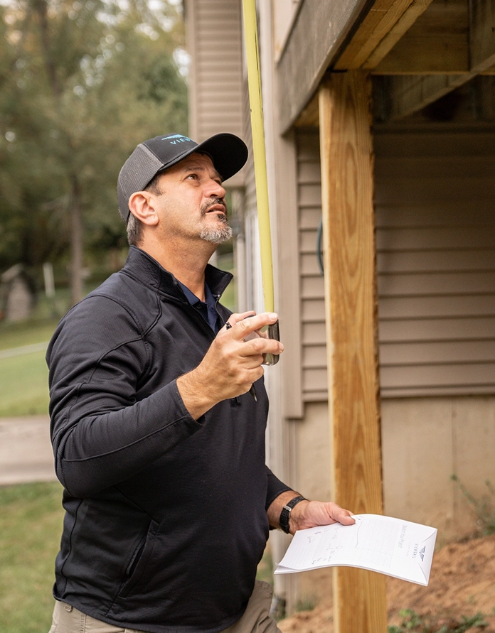 Man in a cap measuring the height of a wooden structure outside a house using a tape measure.