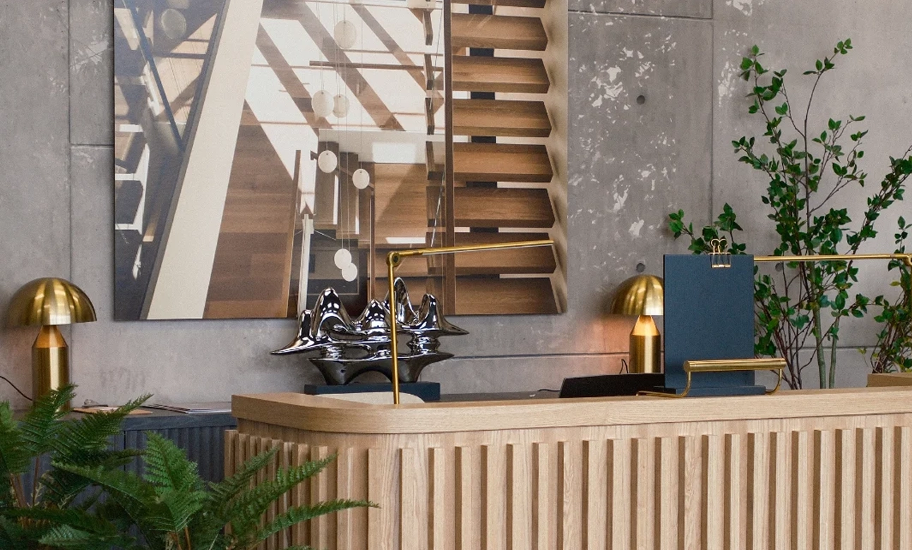 Modern showroom lobby with a wooden reception desk, metallic sculpture, golden lamps, and green plants.
