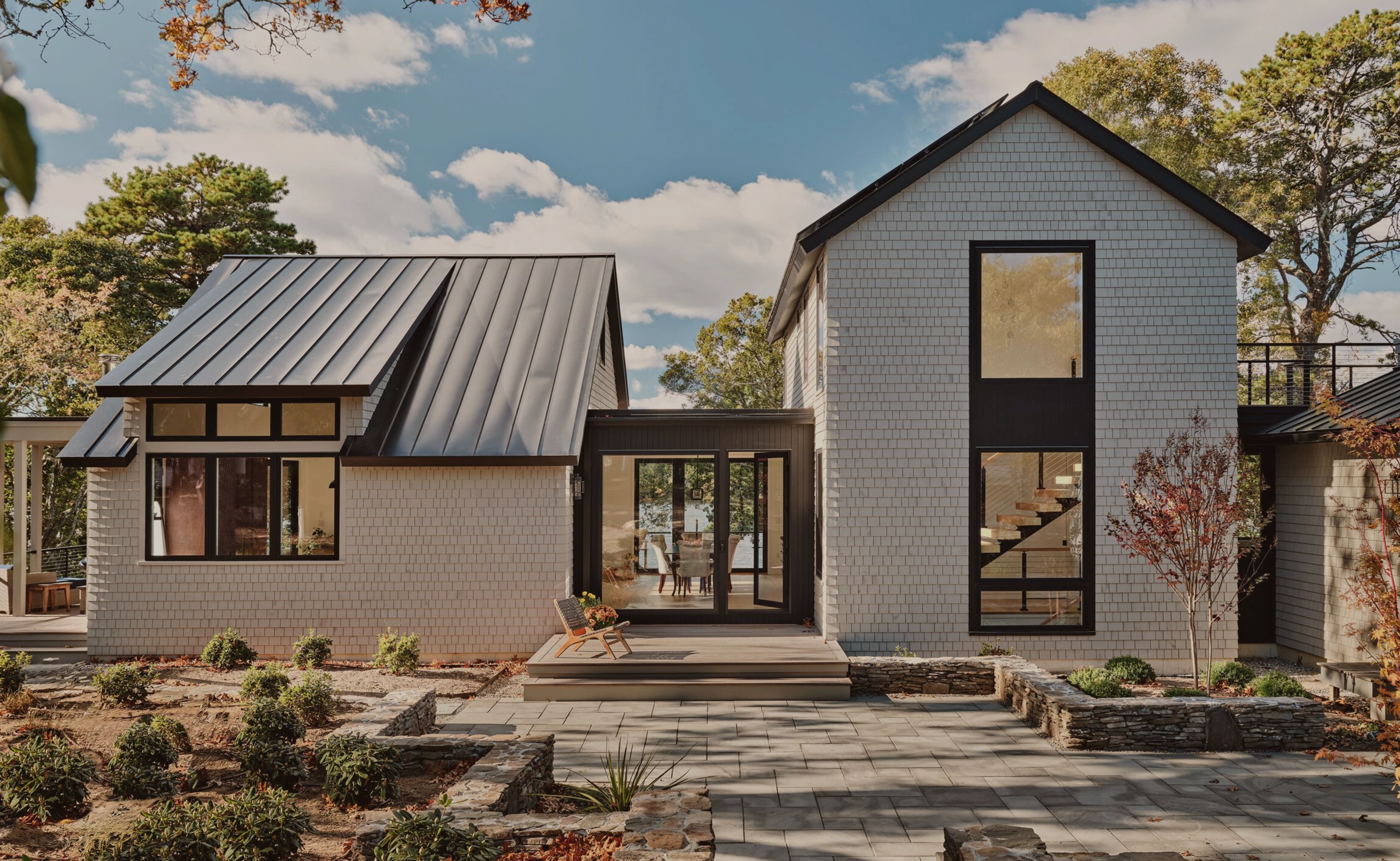 Modern house exterior with light gray shingles, large windows, metal roof, patio, and landscaped garden beds, with floating stairs visible through a two-story window.