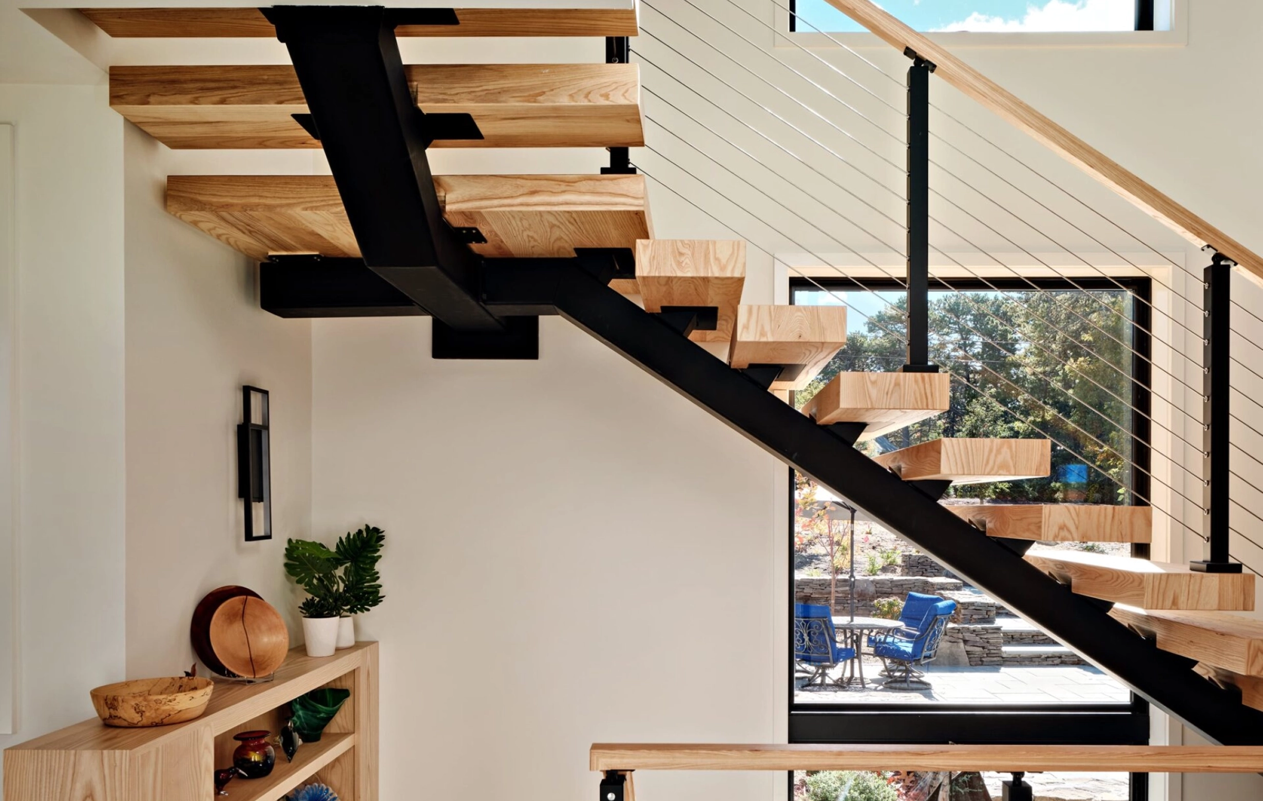 Modern staircase with thick wooden steps and black metal support near a large window showing outdoor patio.