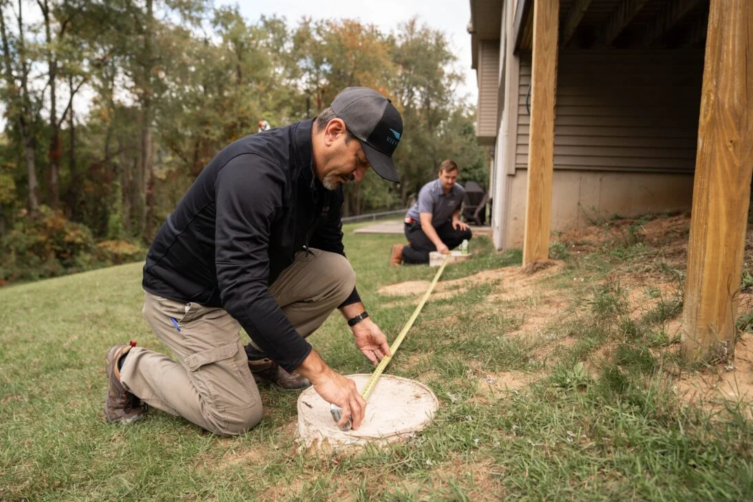 Two installers stretch a tape between concrete footings on a lawn beside a house