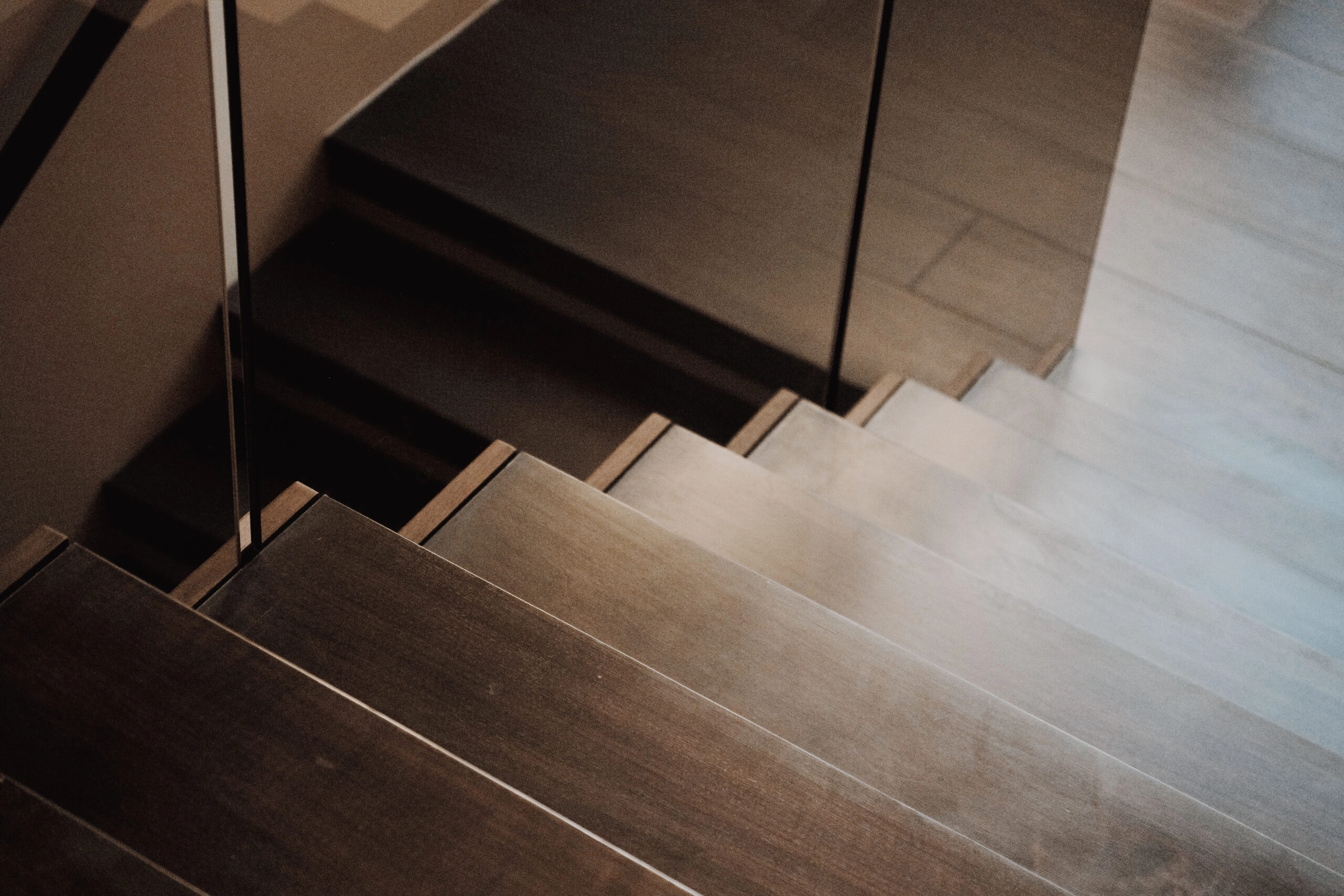 Modern wooden staircase with broad, flat steps and a glass railing reflecting the stairs and surroundings.