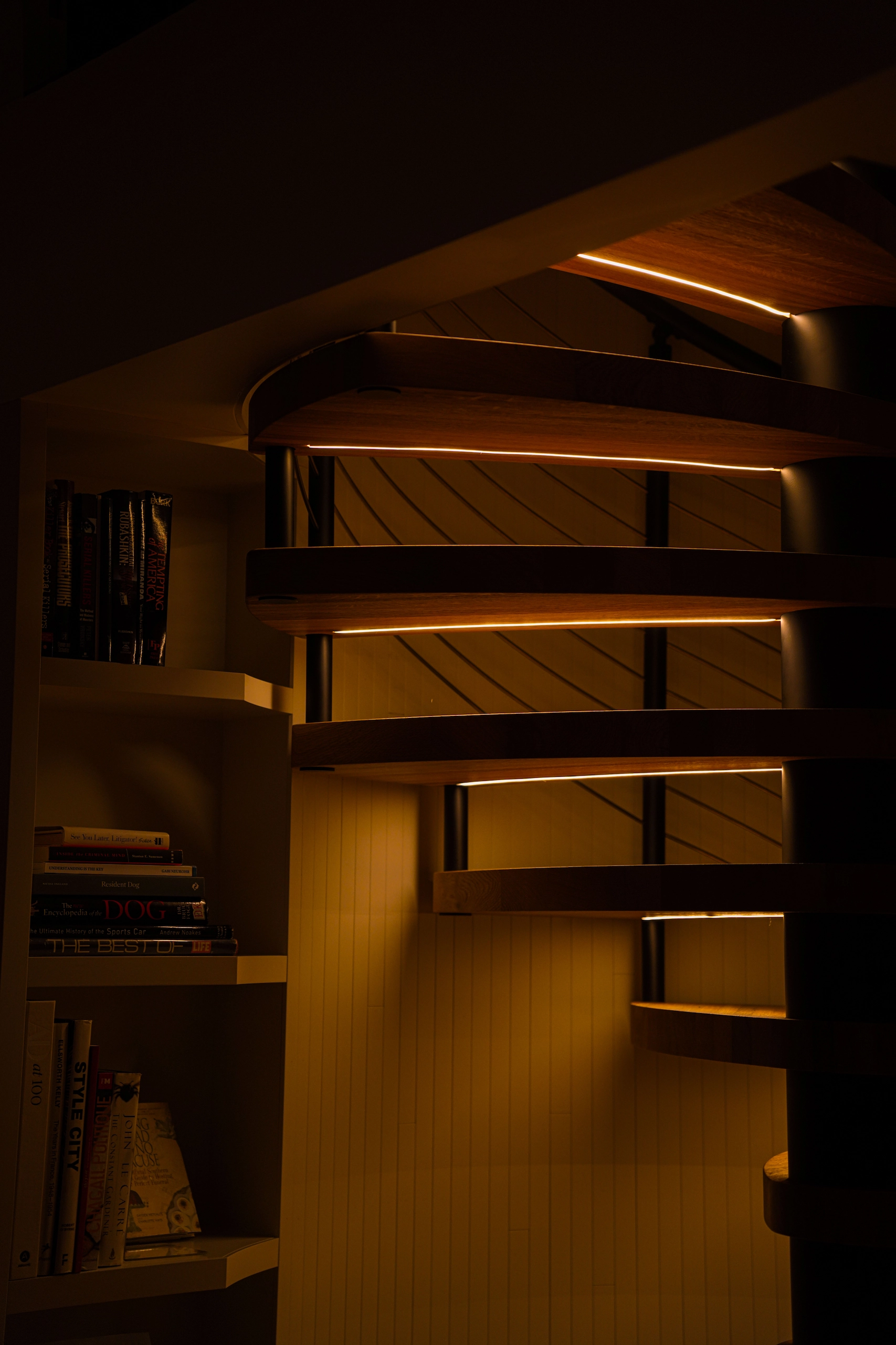 Lit from beneath, each wood tread of a FLIGHT Spiral throws a warm line across the shelves nearby