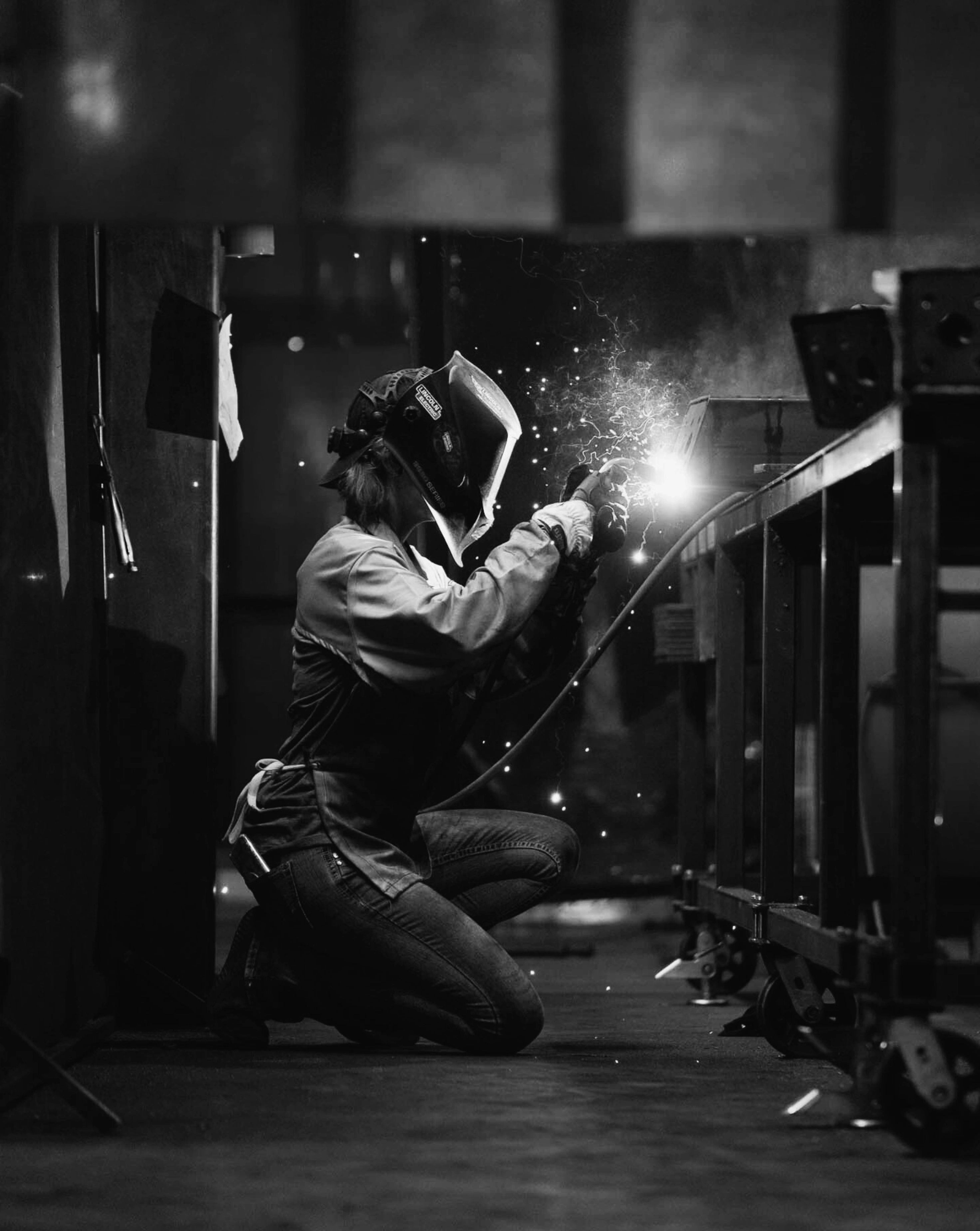 A welder in a protective helmet and gloves kneels while welding metal, with sparks flying off the welding point in a workshop.