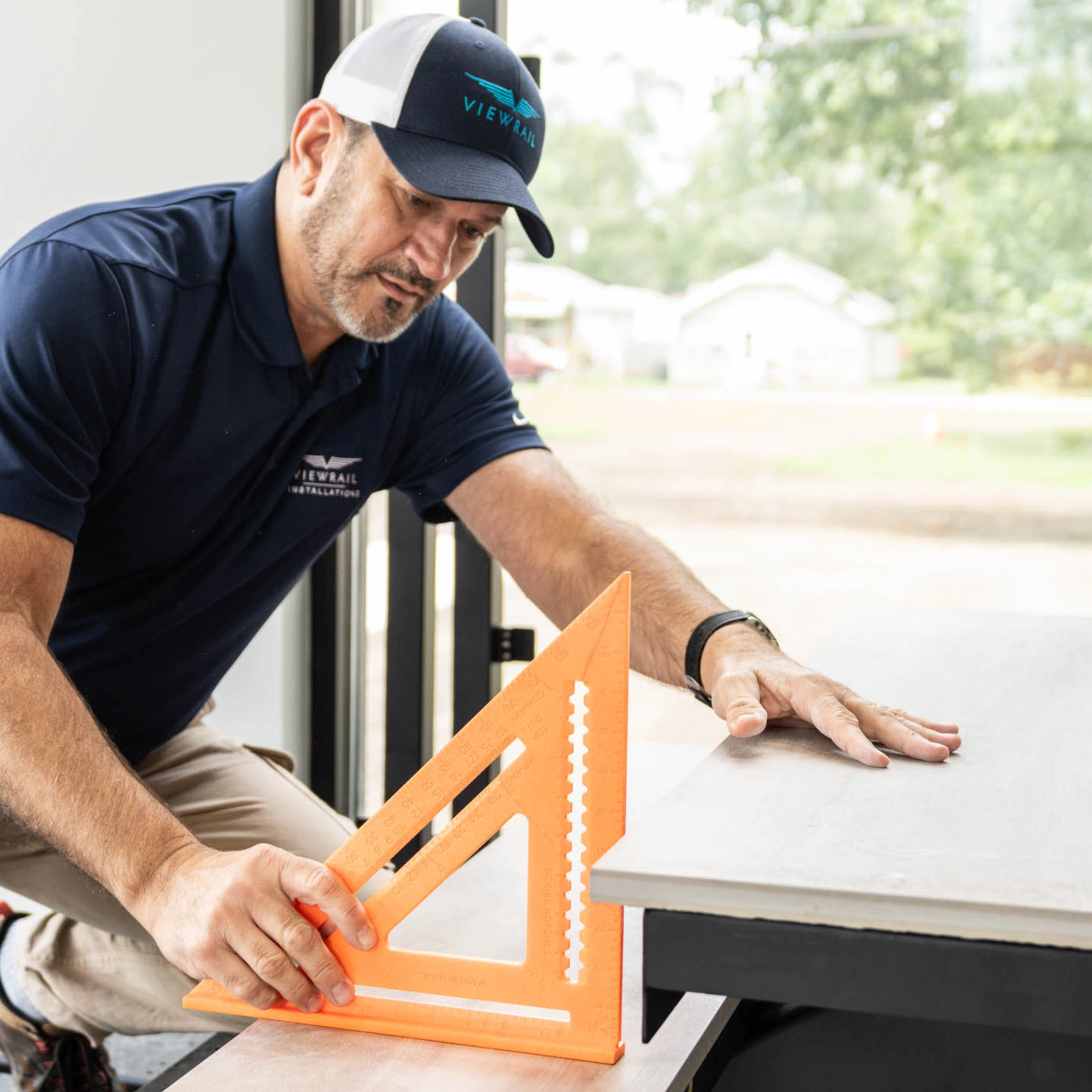 An installer in a Viewrail cap marks a cut with an orange speed square at a saw table