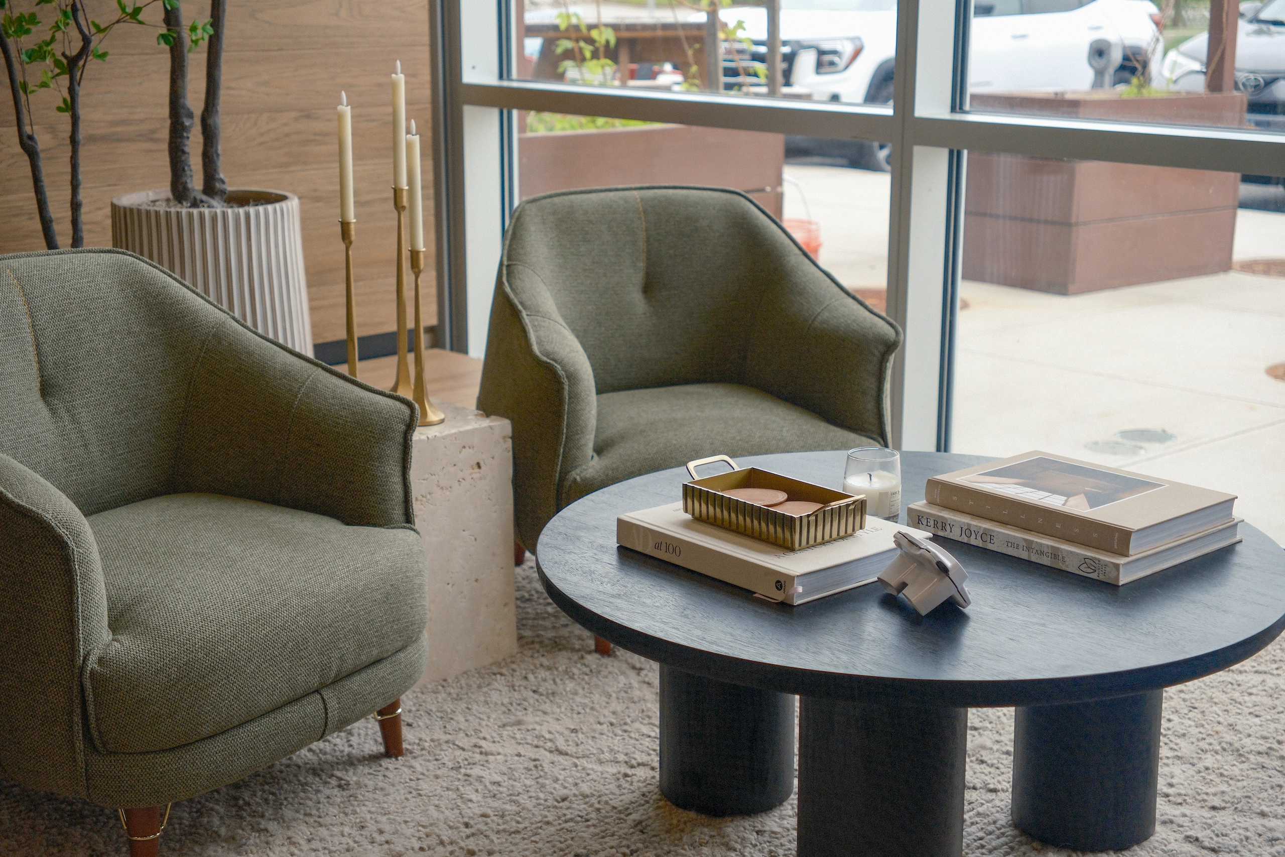 Two green upholstered armchairs around a round black coffee table with books, a small candle, and a brass tray with coasters, set on a beige shag rug in front of large windows.