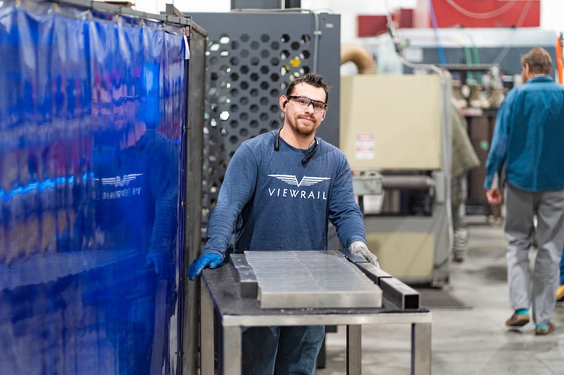 Man wearing safety glasses and gloves standing in an industrial workshop next to a blue barrier and a metal table with rectangular metal pieces, with other workers and machinery visible in the background.