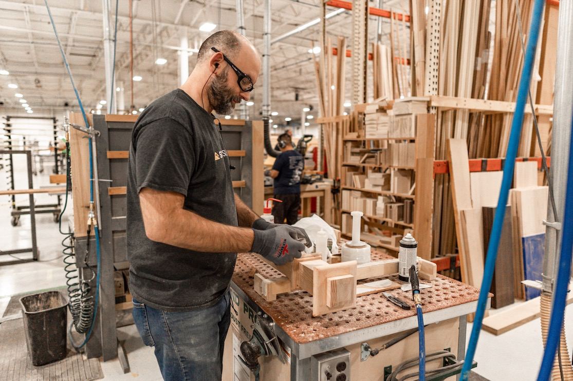 Man wearing safety glasses and gloves working with wooden pieces on a workbench in a busy wood production workshop.