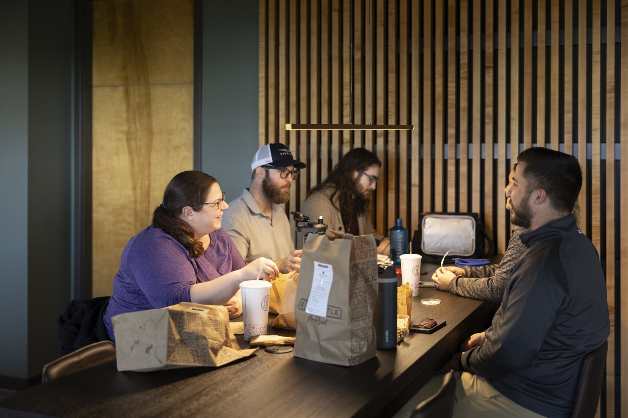 Five people sitting around a wooden table in a modern room with wooden slat walls, eating and talking, with fast food bags, cups, and personal items on the table.