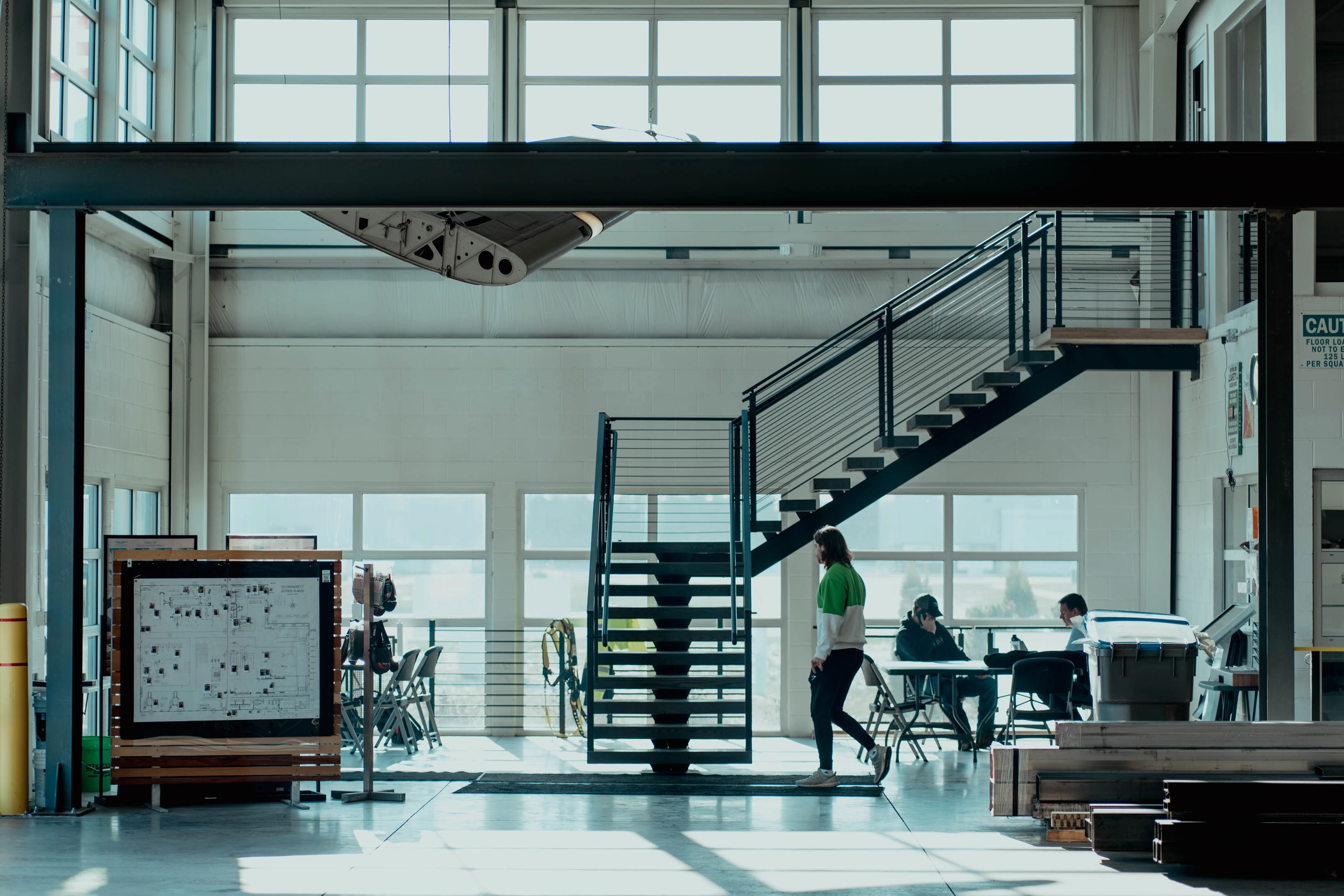 Industrial indoor space with a large metal staircase in the center, two people sitting at a table on the right, and a person in a green and white jacket walking near the stairs, with large windows in the background letting in daylight.