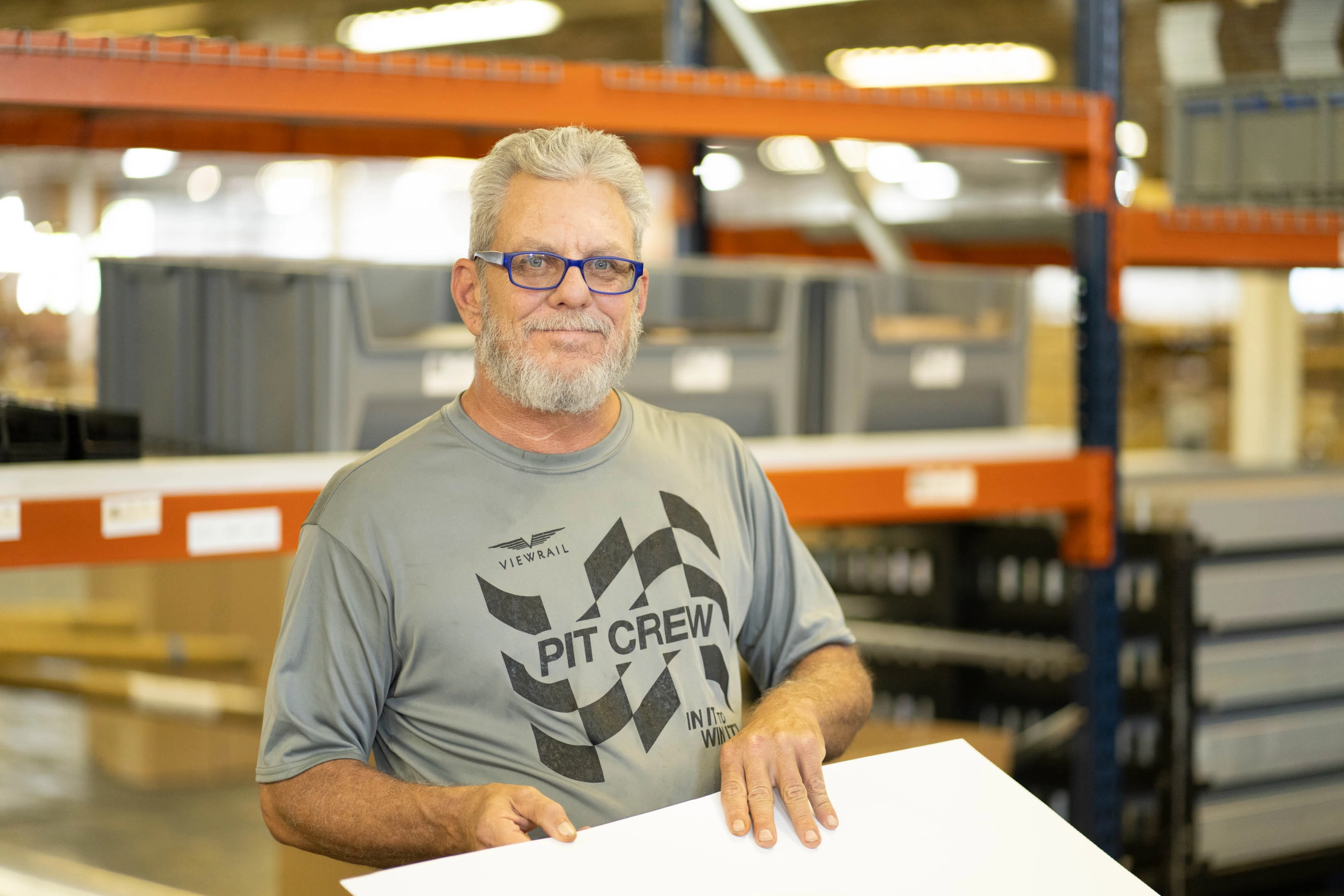 A middle-aged man with gray hair and beard wears blue glasses and a gray 'PIT CREW' t-shirt, holding a white board in a warehouse with shelves and boxes in the background.