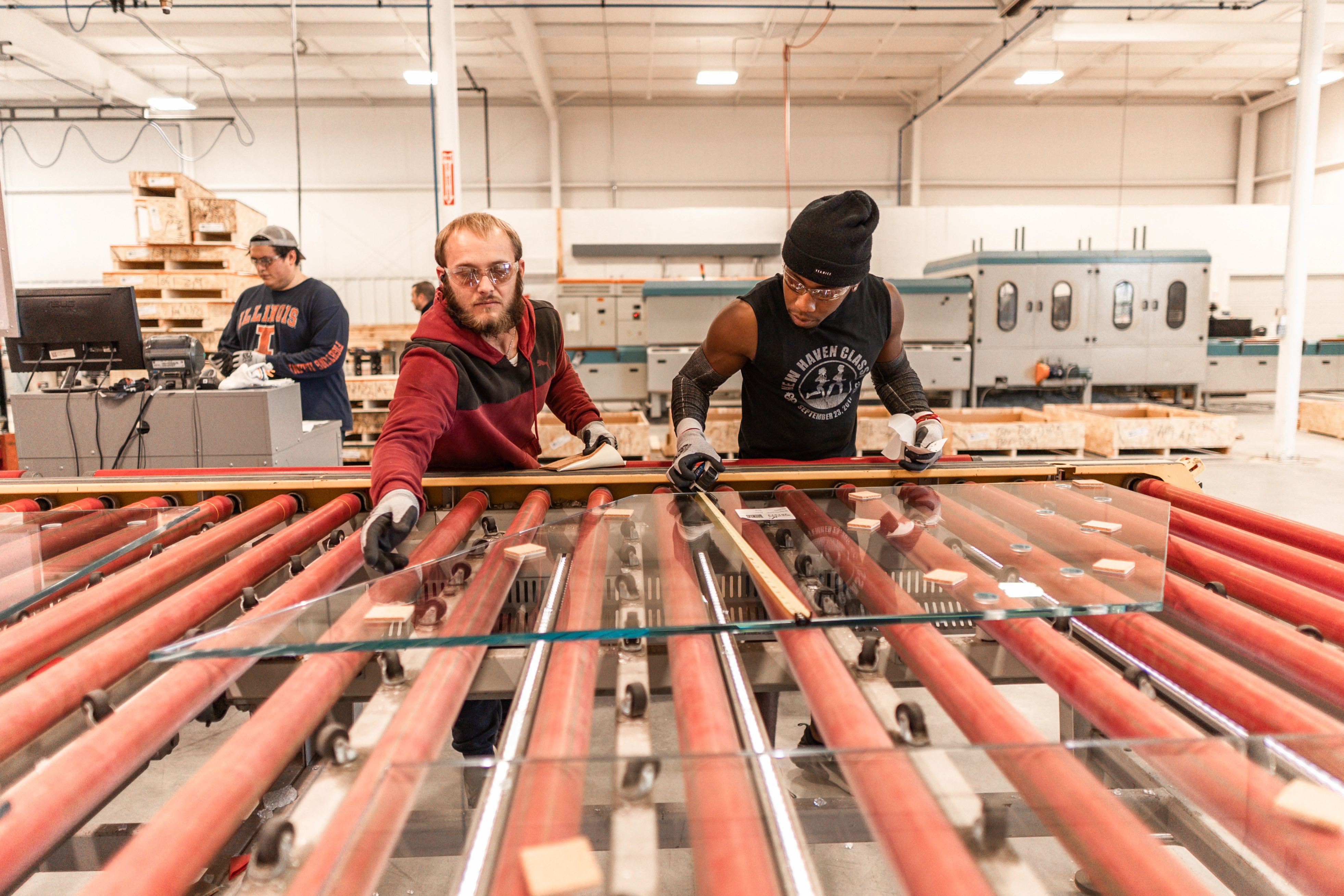 Two workers wearing safety glasses and gloves inspecting large glass sheets on a roller conveyor in a glass production facility, with a third worker operating a computer in the background.