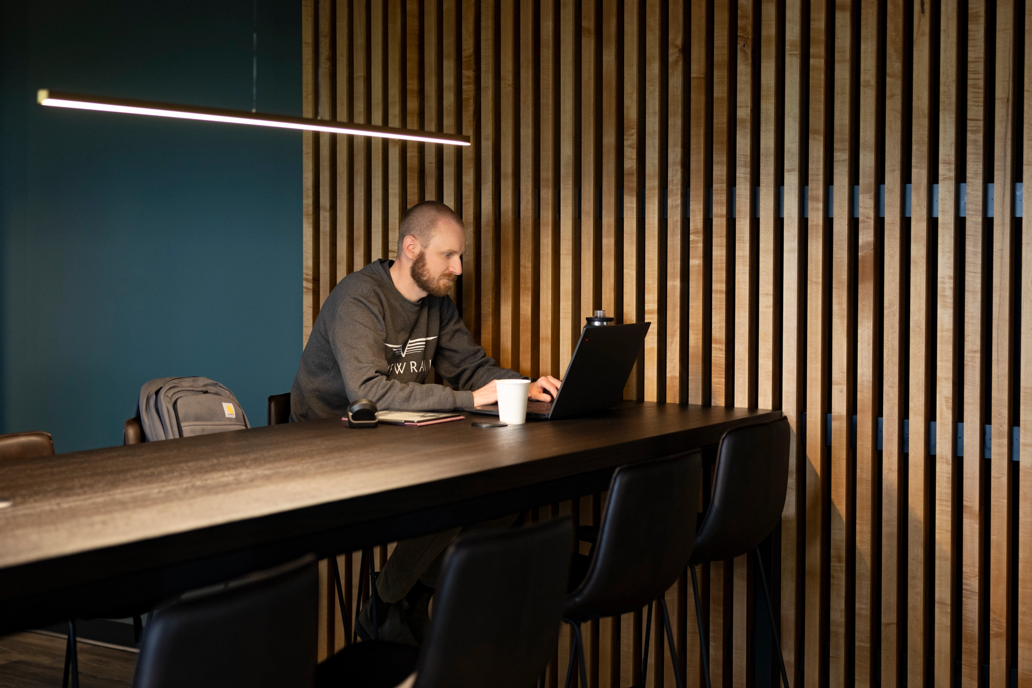 A man with a beard working on a laptop at a long wooden table in a room with vertical wooden slats on the wall and a modern linear light above.