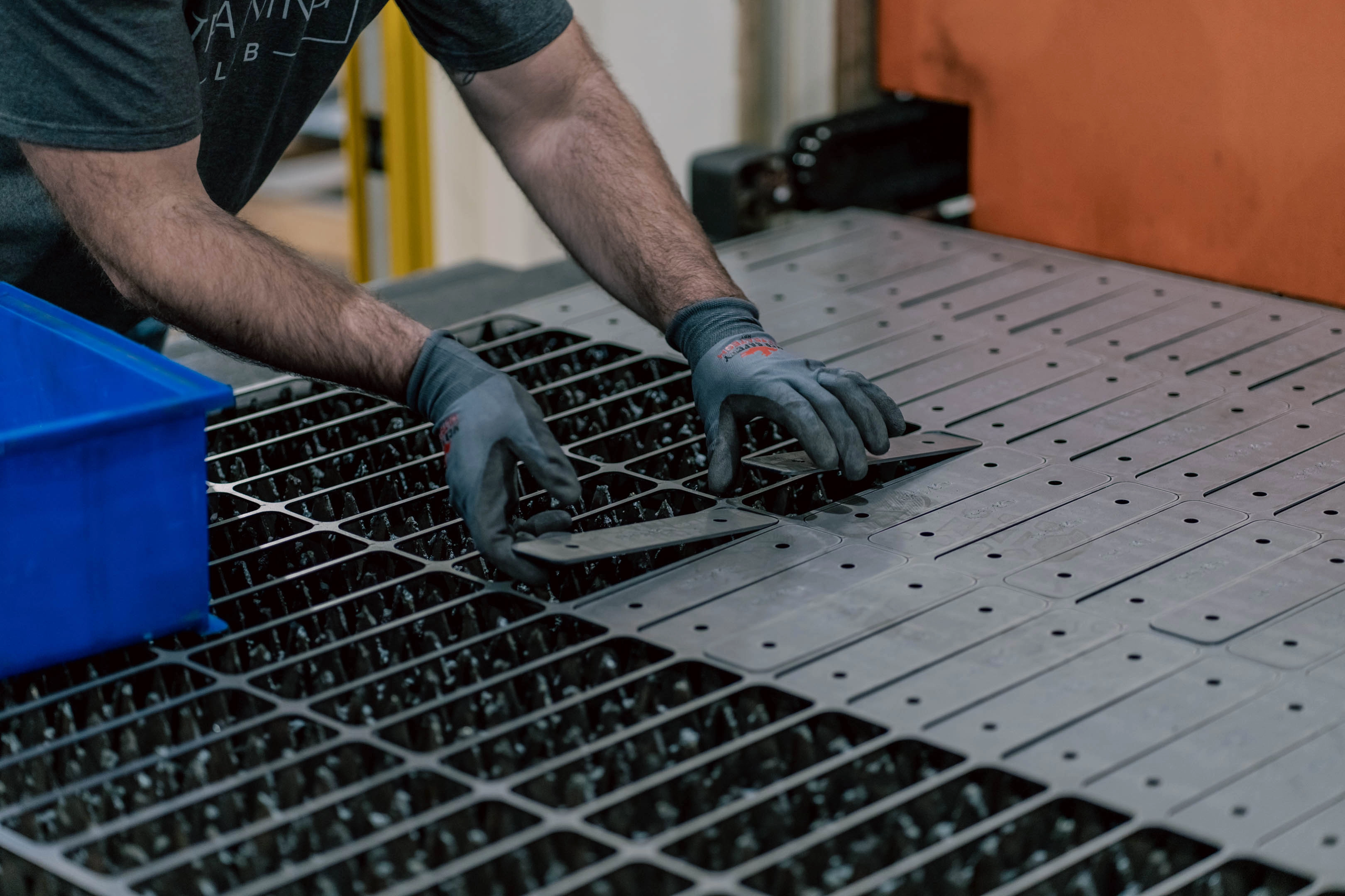 Worker wearing gloves handling metal plates on a factory machine table, arranging rectangular metal parts with holes.